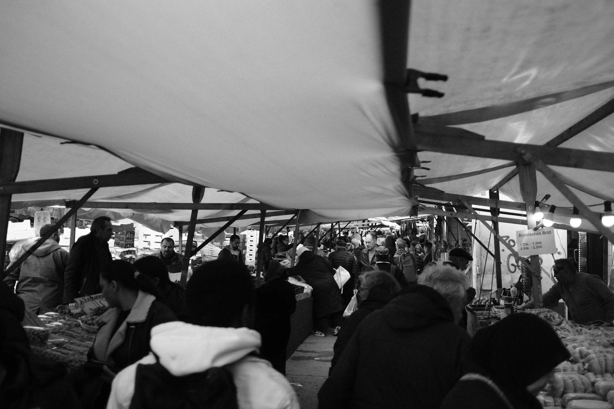 Menschen auf einem Markt, die verschiedene Produkte verkaufen und kaufen, unter Zelten mit Marktschildern in einer belebten Umgebung, schwarz-weiß Fotografie.