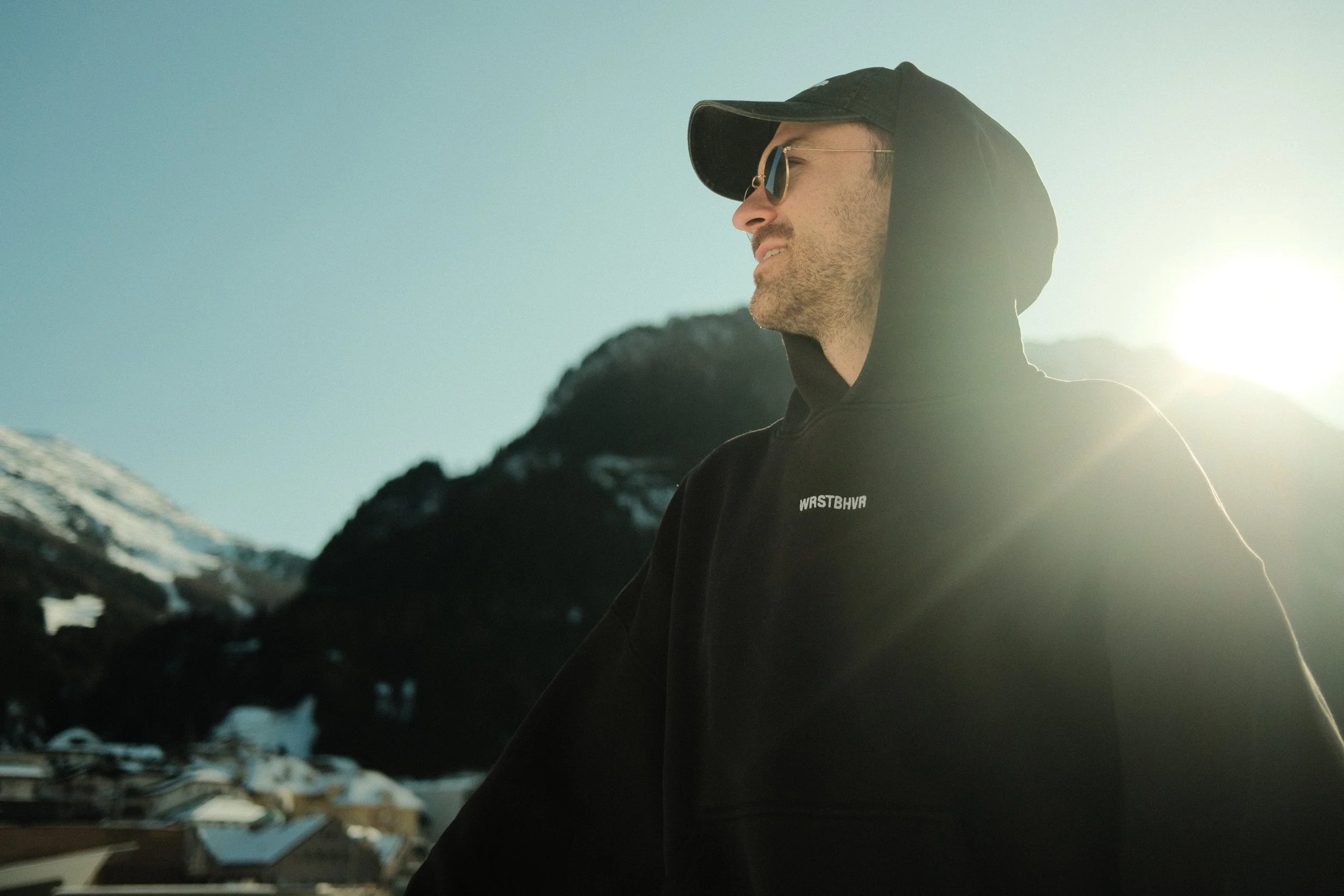 Ein Mann trägt eine schwarze Kapuzenjacke und eine schwarze Baseballkappe, Sonnenbrille, steht vor einer Berglandschaft bei Sonnenaufgang oder Sonnenuntergang.