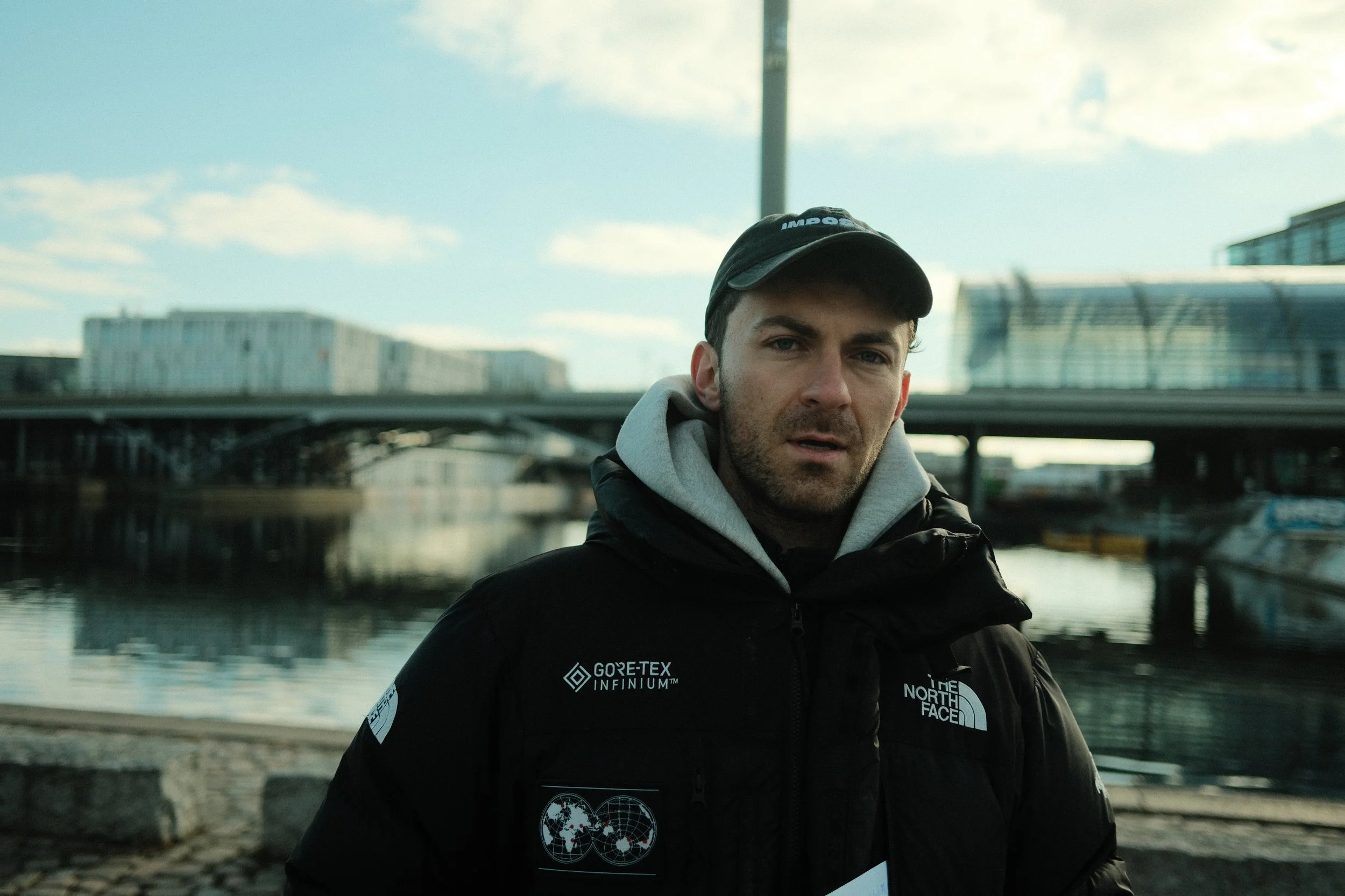 Ein junger Mann trägt eine schwarze Jacke und eine schwarze Kappe, steht am Wasser mit moderner Stadt im Hintergrund, bewölkter Himmel, während die Spiegelung der Stadt im Wasser sichtbar ist.