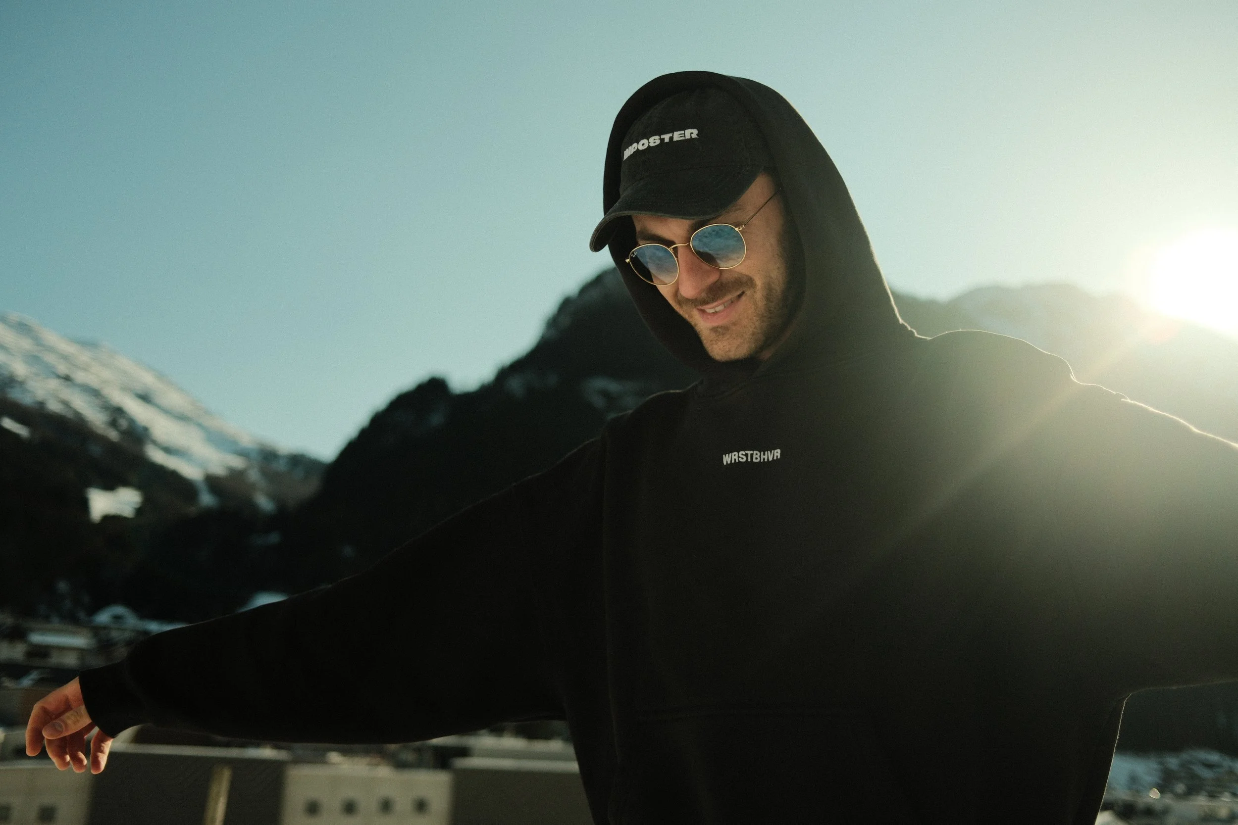 Junge Person trägt schwarze Kapuzenjacke, Baseballkappe und runde Sonnenbrille, Sonnenlicht im Hintergrund.