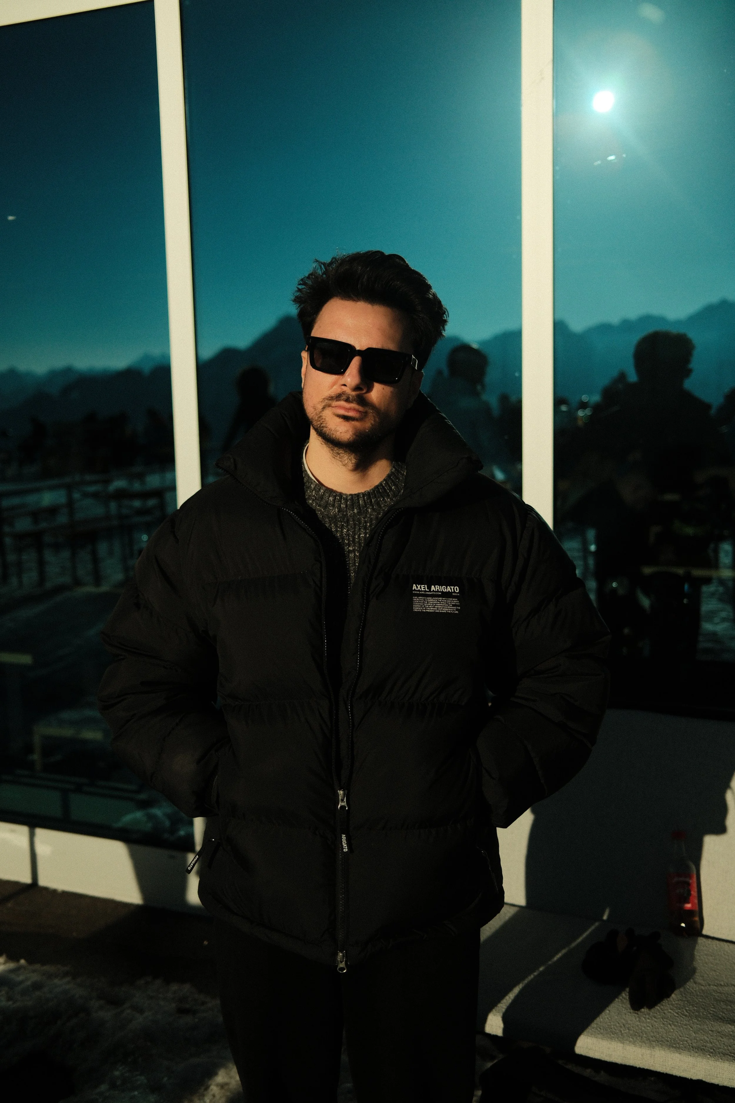 Ein Mann trägt schwarze Sonnenbrille und schwarze Jacke, steht vor einer reflektierenden Wand mit Bergkulisse bei Nacht.