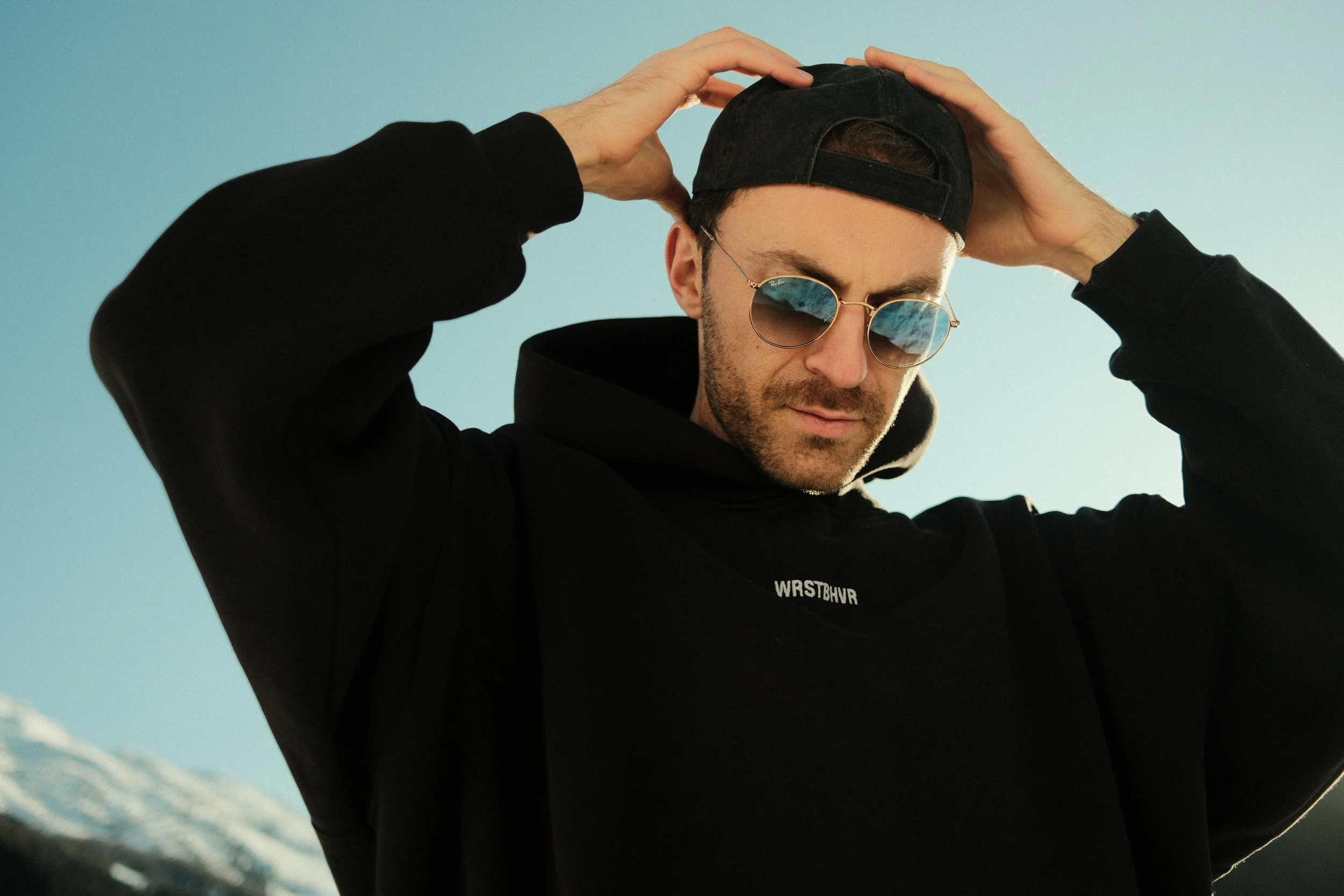 Junge Person trägt schwarze Kapuzenjacke, Sonnenbrille und schwarze Cap, sieht nach unten, Hintergrund blau, evtl. Himmel.