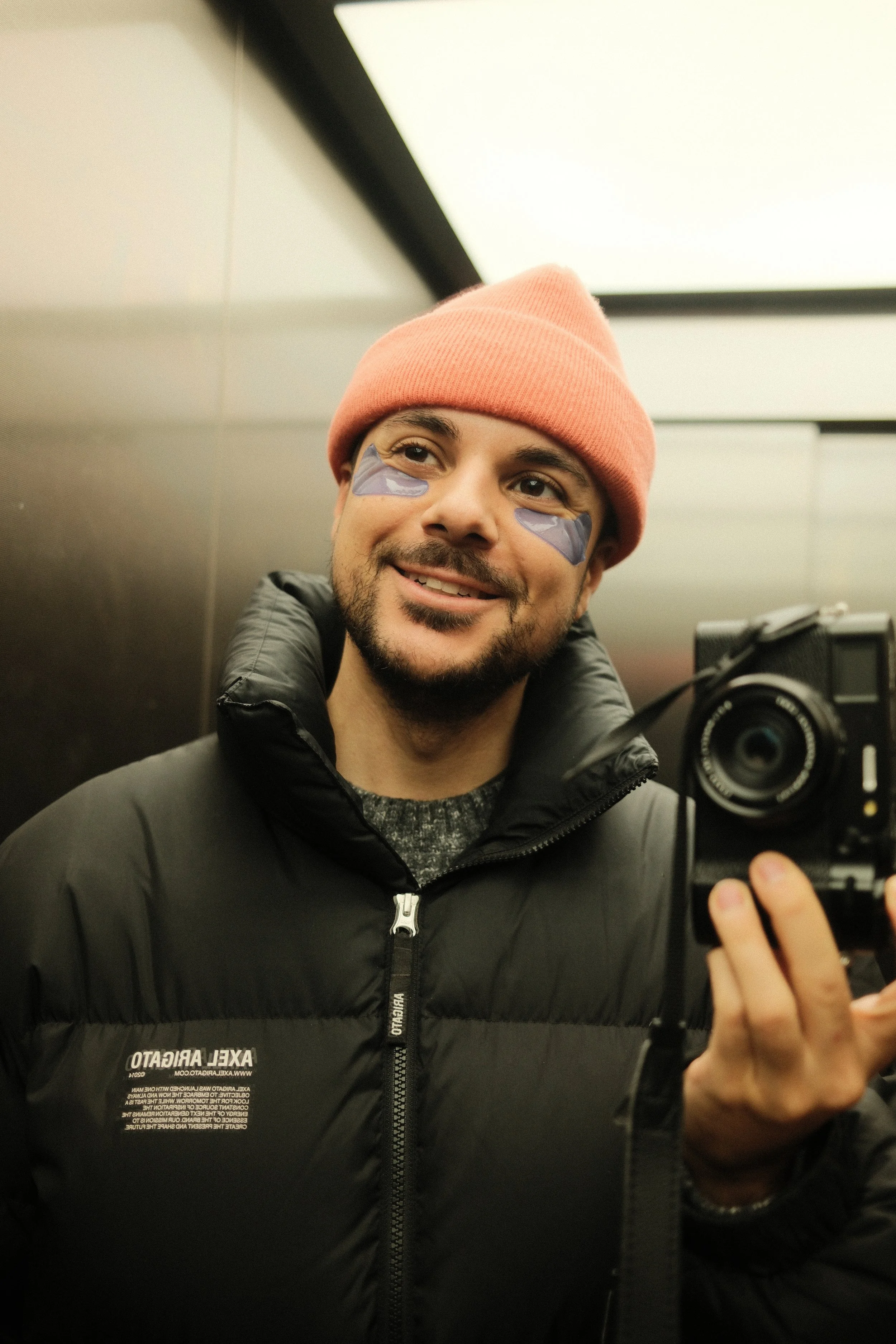 Ein Mann trägt einen pinkfarbenen Beany-Mütze und eine schwarze Jacke, steht in einem Aufzug und nimmt ein Selfie mit einer Kamera. Der Mann hat einen Bart und hat unter den Augen Make-up.