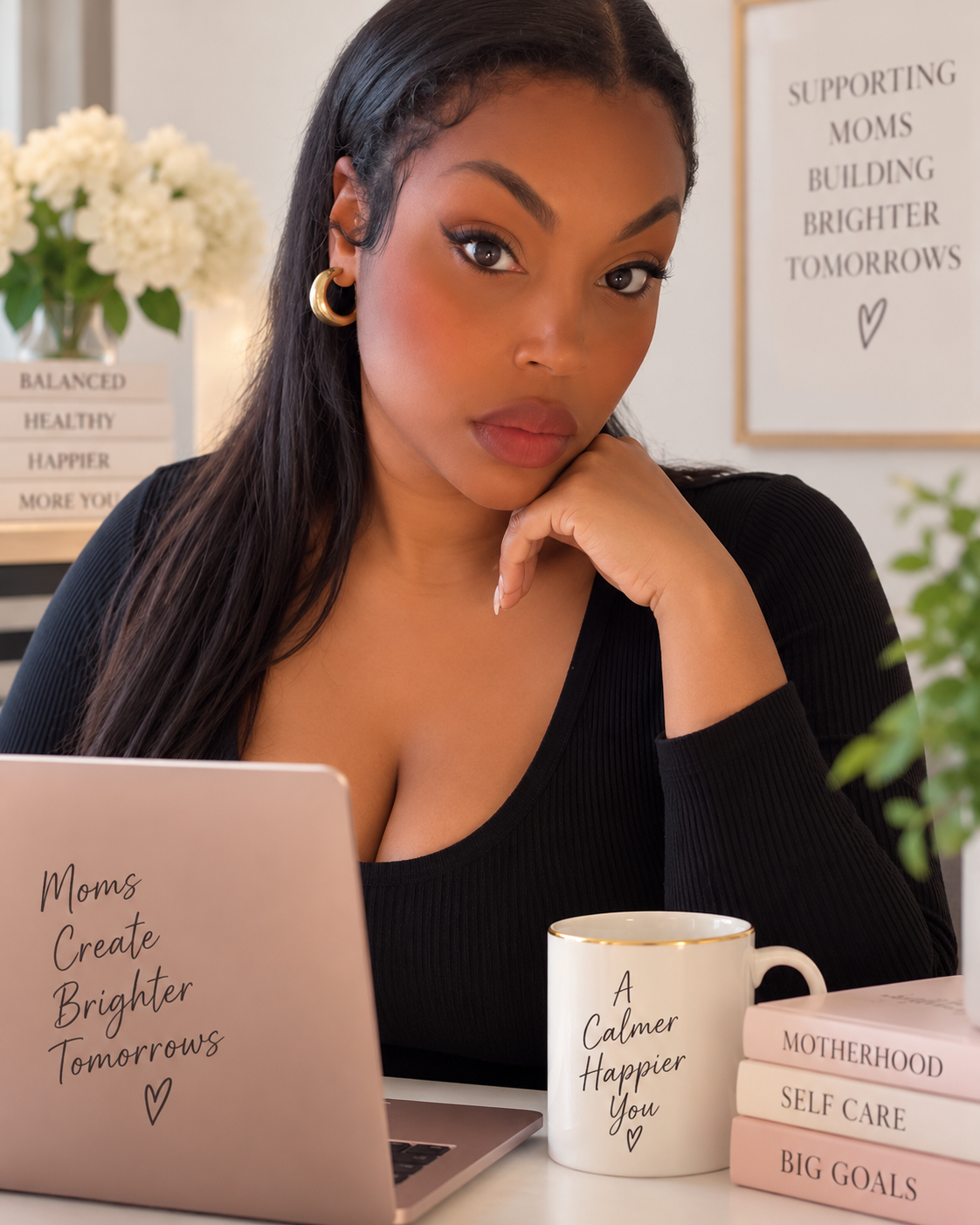 A woman sitting at a desk, looking towards the camera, with a laptop, a white mug with the message "A Calmer Happier You," a stack of pink books titled "Motherhood," "Self Care," "Big Goals," and "Self Love," and a background sign that reads "Supporting Moms Building Brighter Tomorrows."