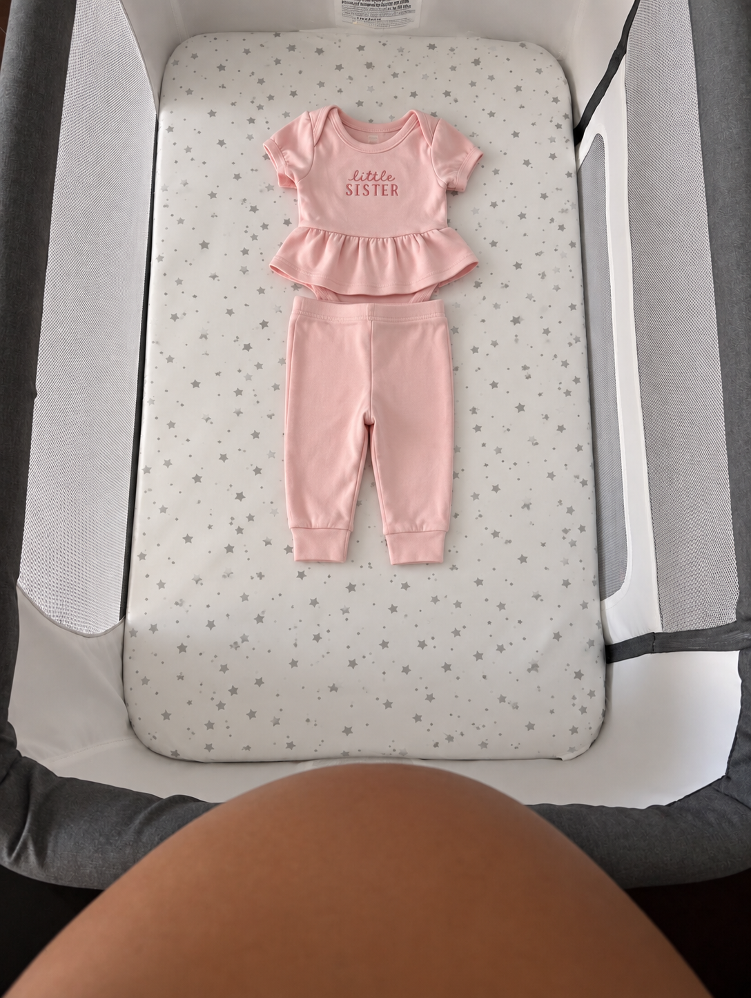 Pink baby outfit with a top that says 'little SISTER' and matching pants on a star-patterned mattress inside a crib.
