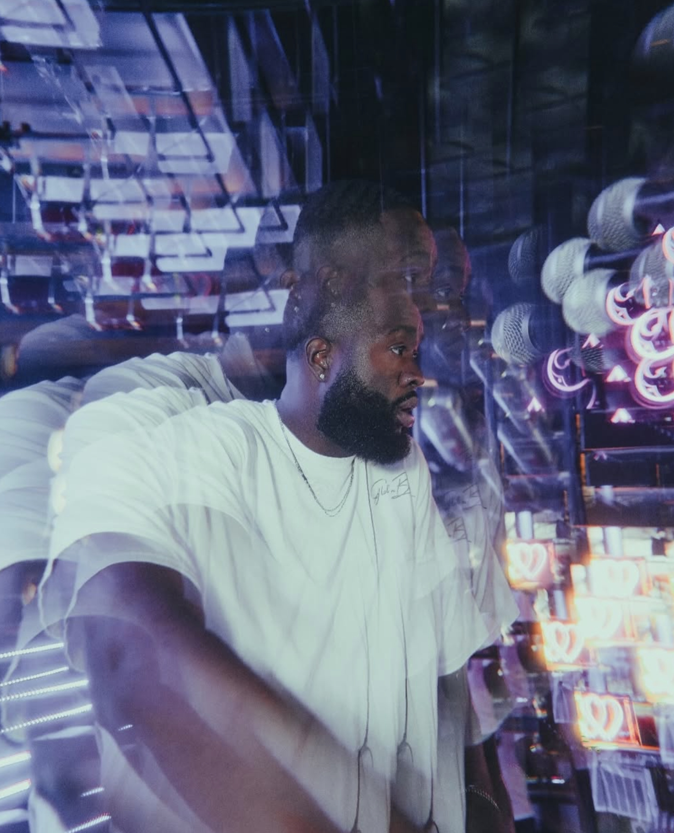 dj-ambitious-boy- with a beard and earrings is looking at himself in a reflective surface at a mirror, surrounded by neon lights and microphone props.