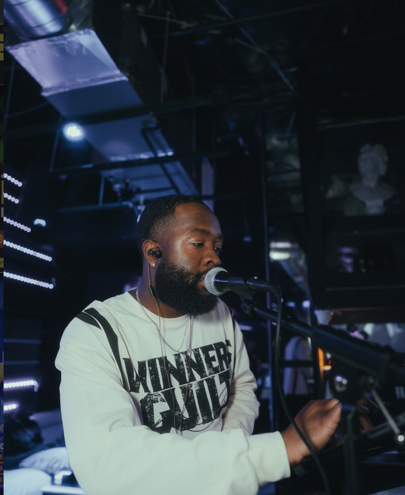 DJ-ambitious-boy, with a beard, wearing a white long-sleeve shirt with black text, is speaking or singing into a microphone in a dimly lit room with blue and purple lighting.