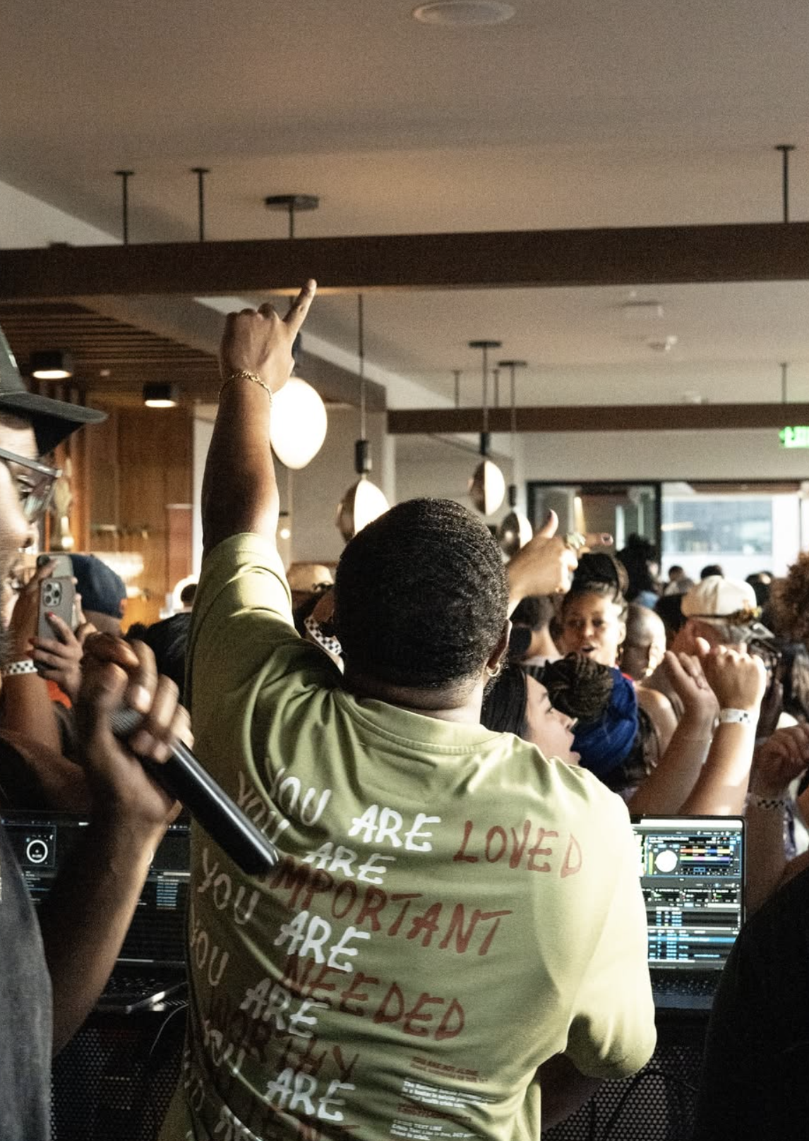 A crowded indoor event with people dancing and taking photos. dj-ambitious-boy- in a yellow shirt with red and white text stands in the foreground, raising their hand. Another person with a microphone is in front of DJ equipment.
