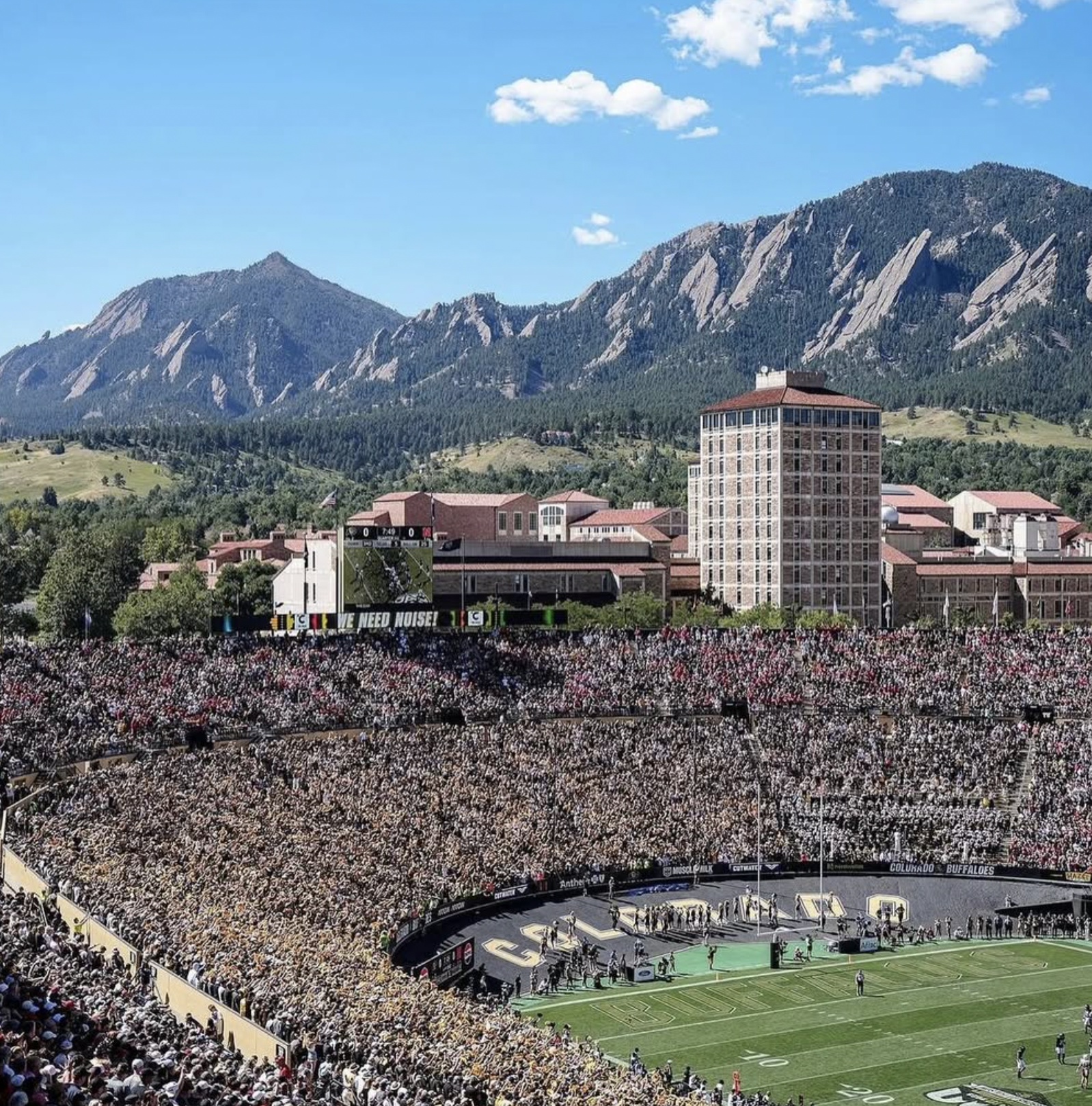 A packed football stadium with a large crowd, green field, and a large screen displaying the score, with mountains and blue sky in the background.