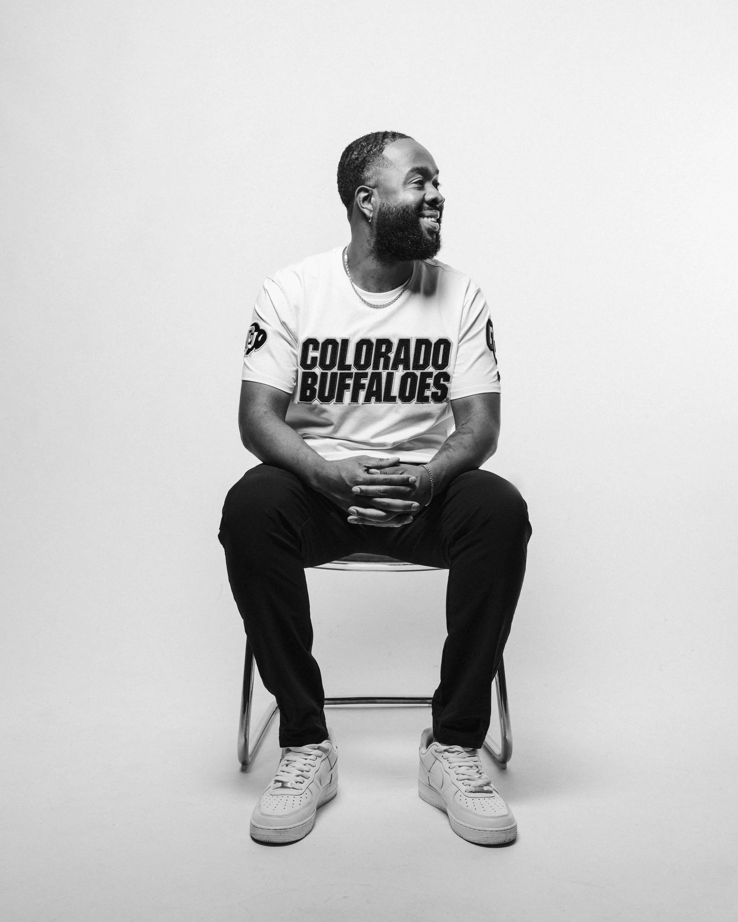 dj-ambitious-boy- with a beard wearing a Colorado Buffaloes shirt, sitting on a stool with hands clasped, smiling and looking to his right, in a black and white photo.