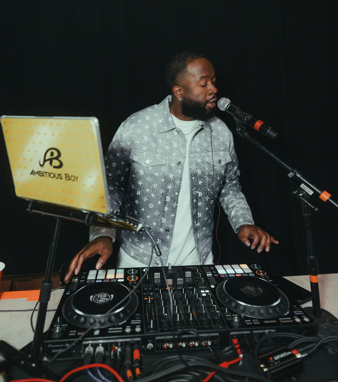 dj-ambitious-boy- performing with a DJ controller, a microphone, and a laptop labeled 'Ambitious Boy' on a stand, set against a black background.