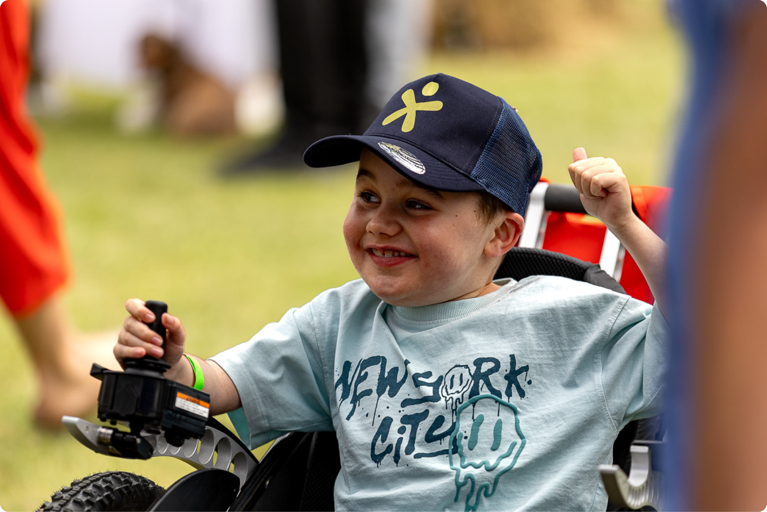 A young boy smiling and raising his fist with a clenched fist, wearing a navy blue cap and a light gray T-shirt with graphics. He is sitting in a wheelchair at an outdoor event on a grassy field, with blurred people in the background.