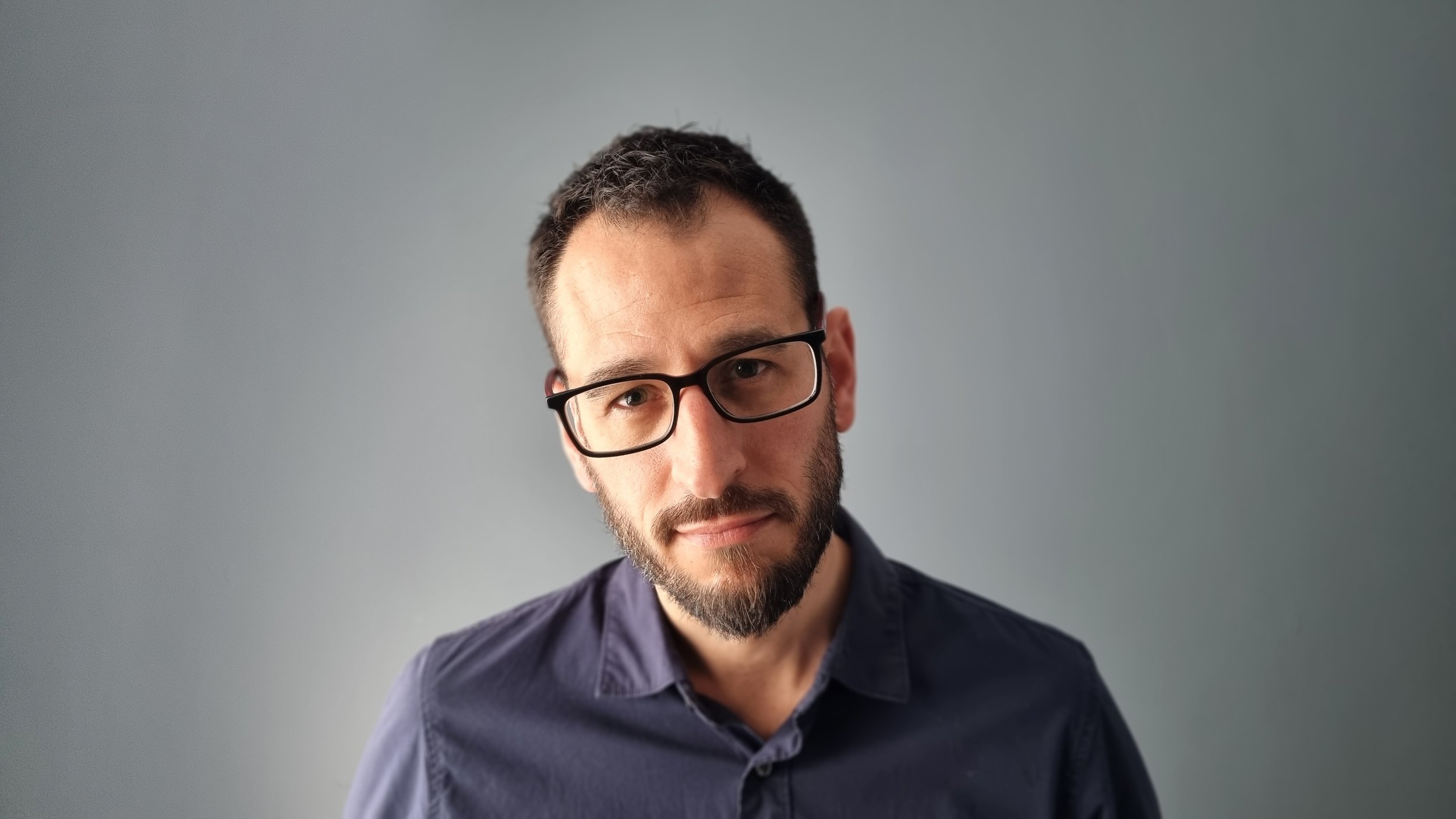 A man with dark hair, glasses, and a beard wearing a dark blue shirt poses against a plain gray background.