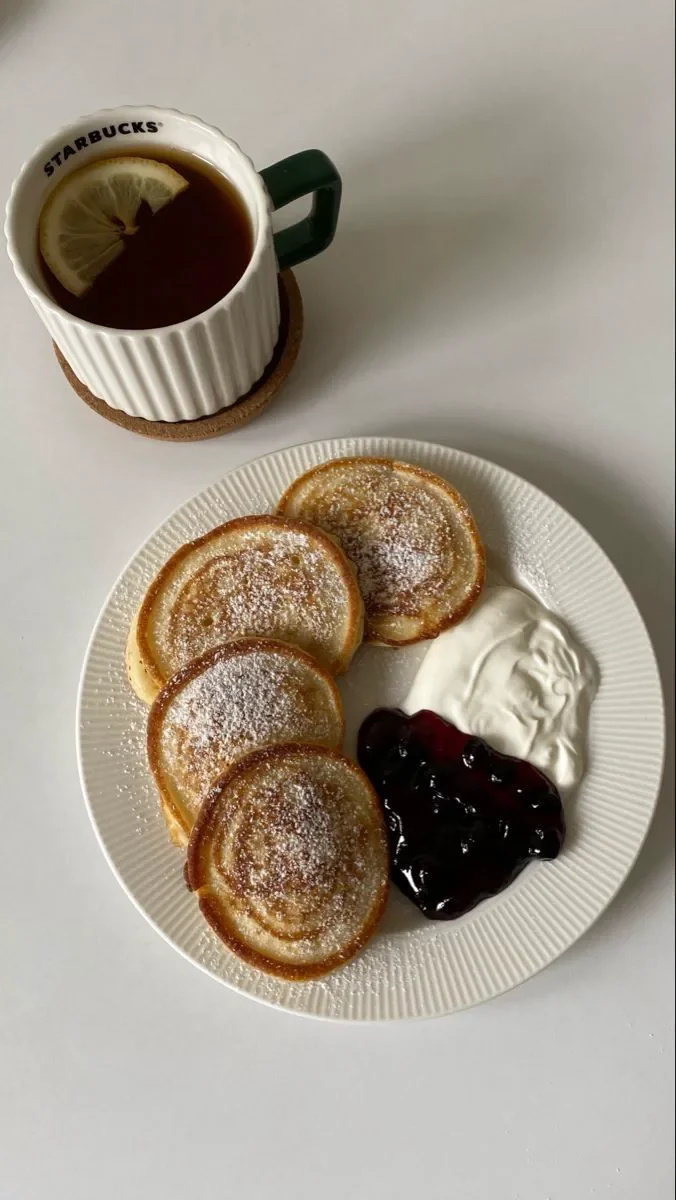 Piatto con quattro crepes dolci, panna montata e marmellata di mirtilli, accompagnato da una tazza di tè con fetta di limone, su un piatto di ceramica bianca
