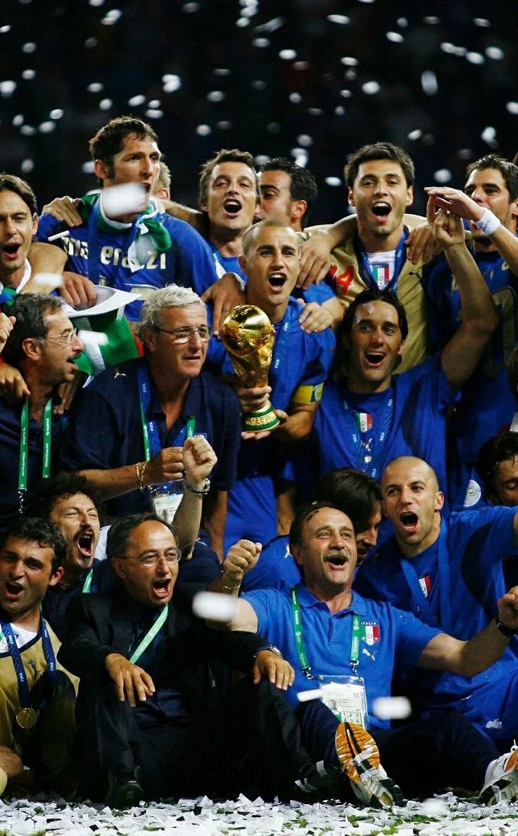 Una squadra di calcio italiana festeggia con il trofeo della Coppa del Mondo, con il gruppo che brinda e si abbraccia, circondata da coriandoli bianchi.
