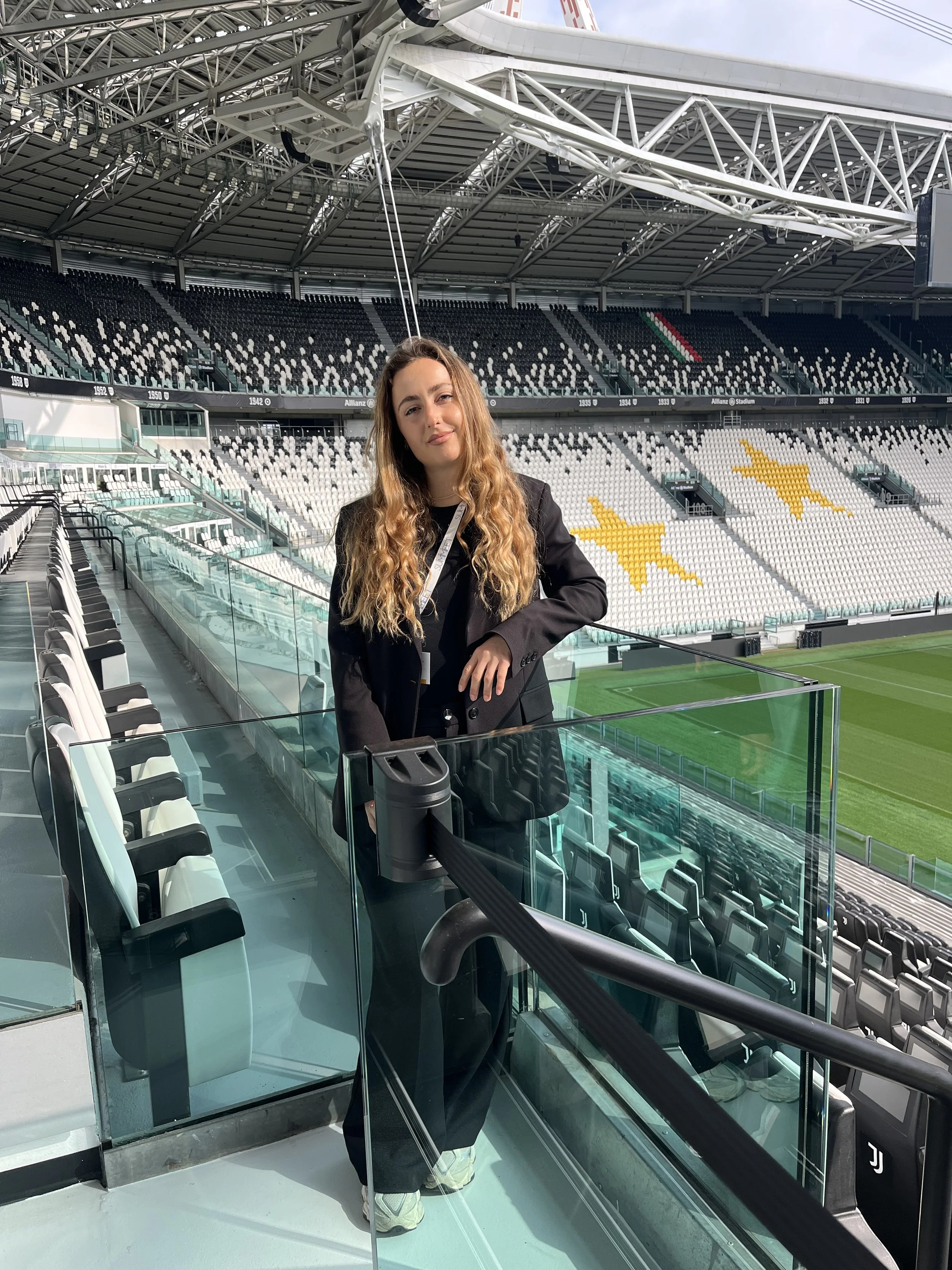 Una donna con capelli lunghi riccioli castani snodati, indossa un abbigliamento nero, si trova all'interno di uno stadio vuoto, davanti a un campo da calcio. La donna sorride con aria tranquilla.