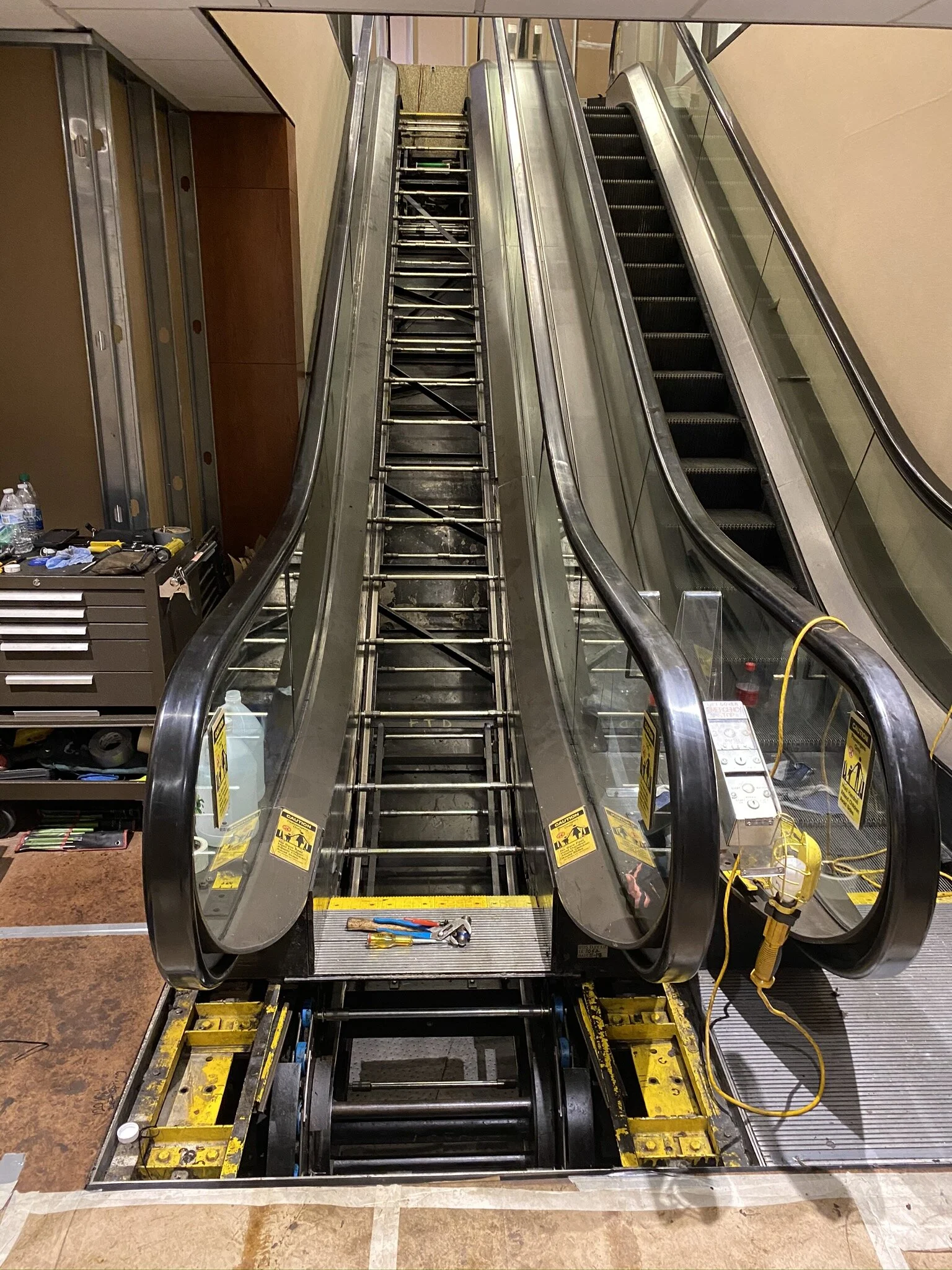 Photo of a disassembled escalator in a building under maintenance. The escalator is missing its steps, and tools are placed on the steps in front of it. The surrounding area includes a brown carpeted floor, a tool chest, and some water bottles.