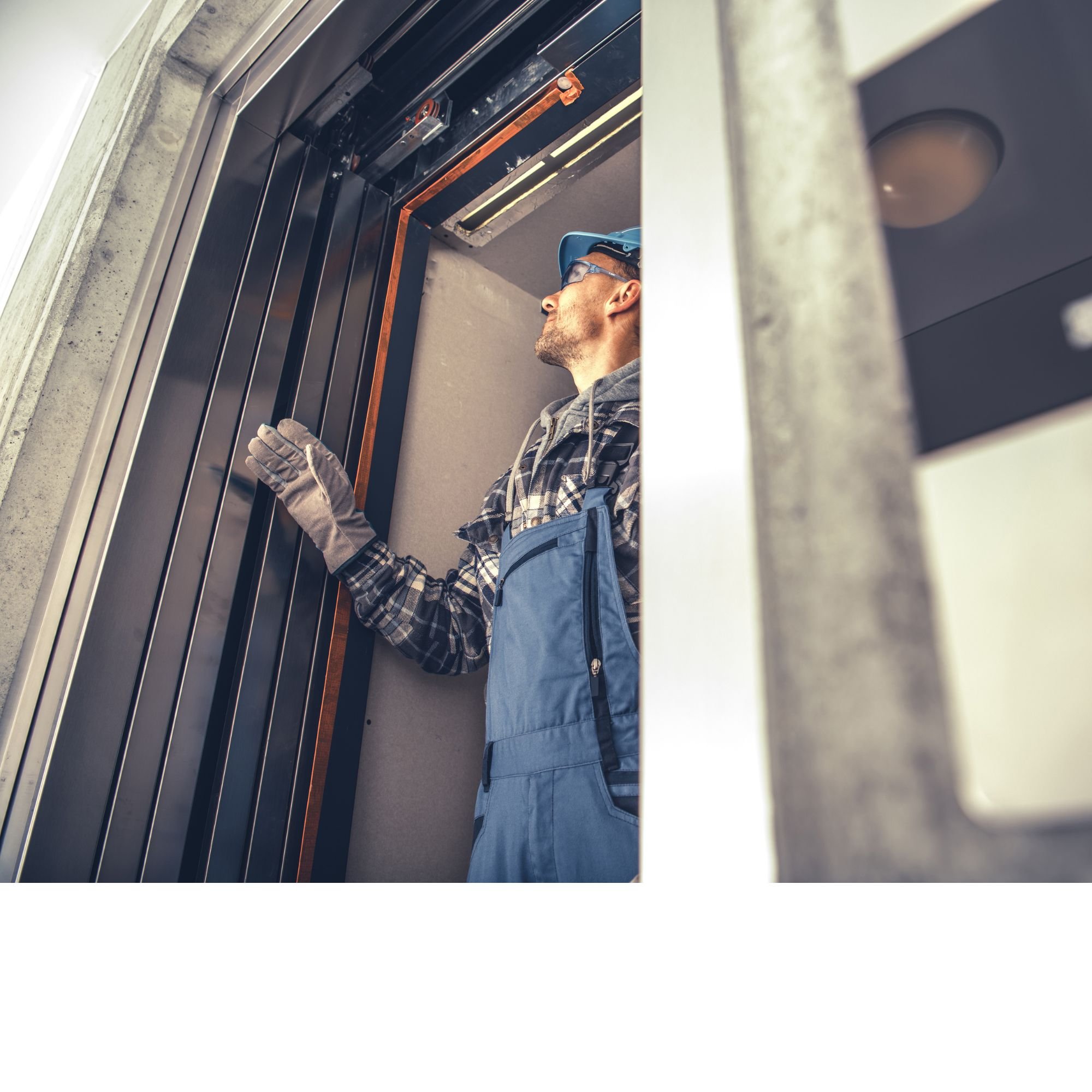 A construction worker wearing a blue hard hat, safety glasses, gray gloves, a plaid shirt, and a gray jacket is working inside a modern elevator shaft with visible metal components and concrete walls.