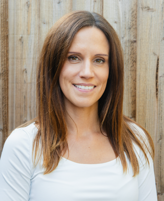 A woman with long, brown hair smiling at the camera, standing in front of a wooden fence.