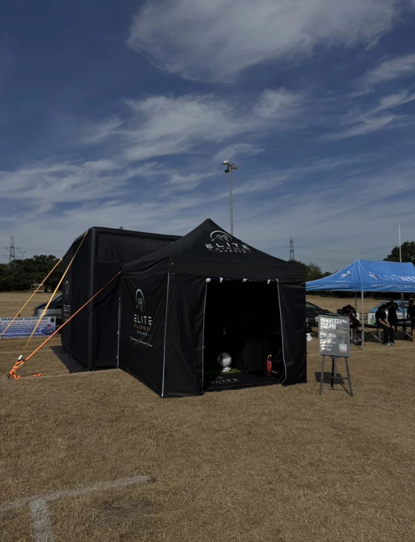 Outdoor event with tents, including a black tent branded 'Elite Fairway,' and a blue tent with people sitting and standing nearby, under a partly cloudy sky.