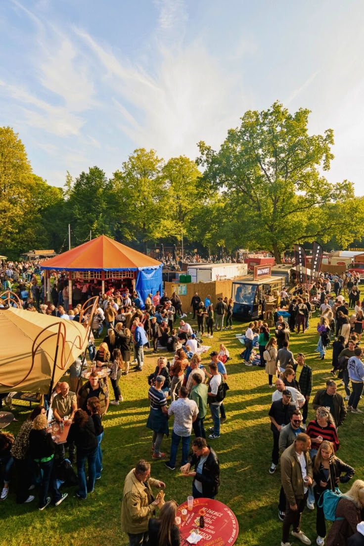 People attending a fair or outdoor event with food stalls, tents, and a large crowd on a grassy area.