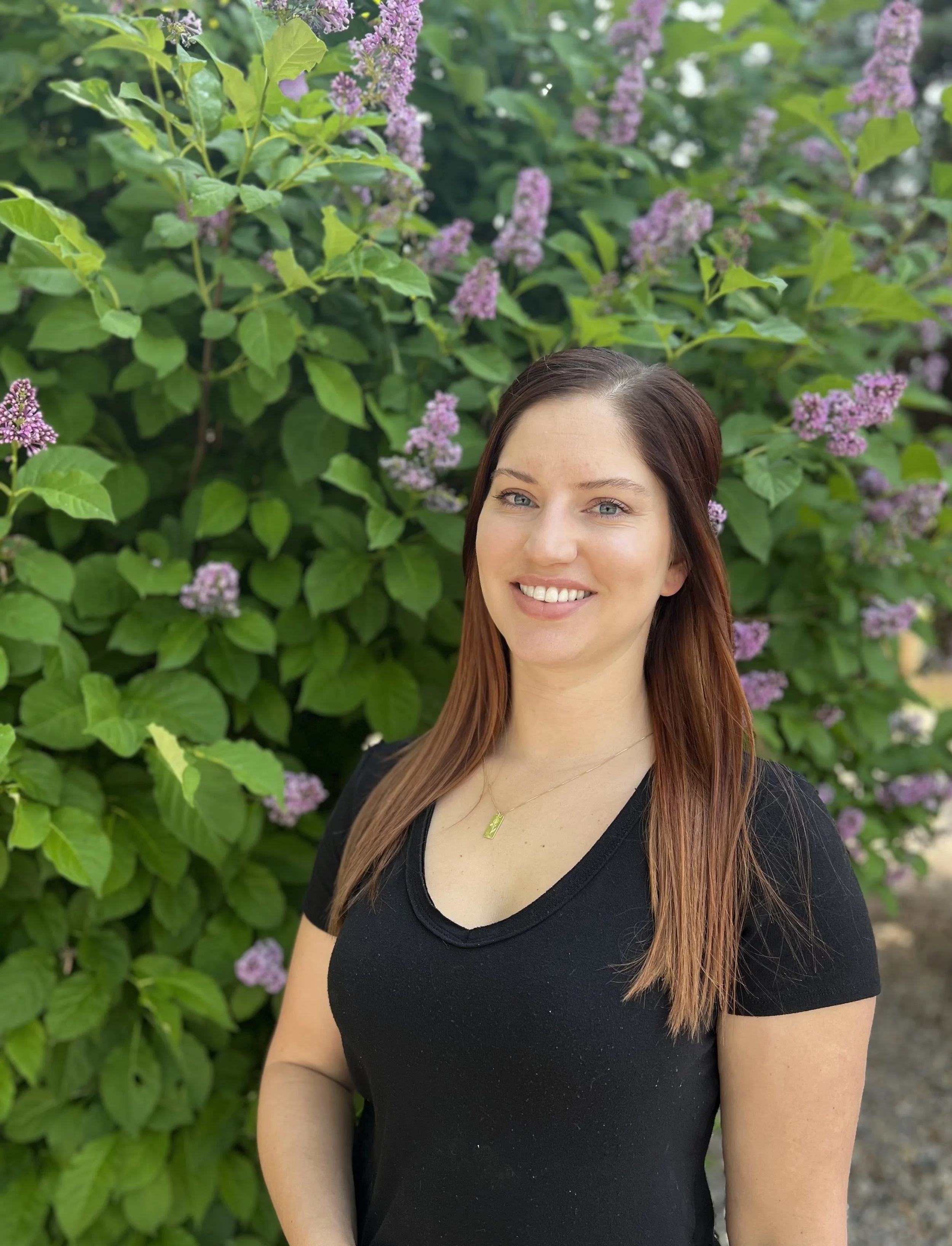 A woman with long, straight, auburn hair and blue eyes, smiling, standing in front of a lush green shrub with pinkish-purple flowers.