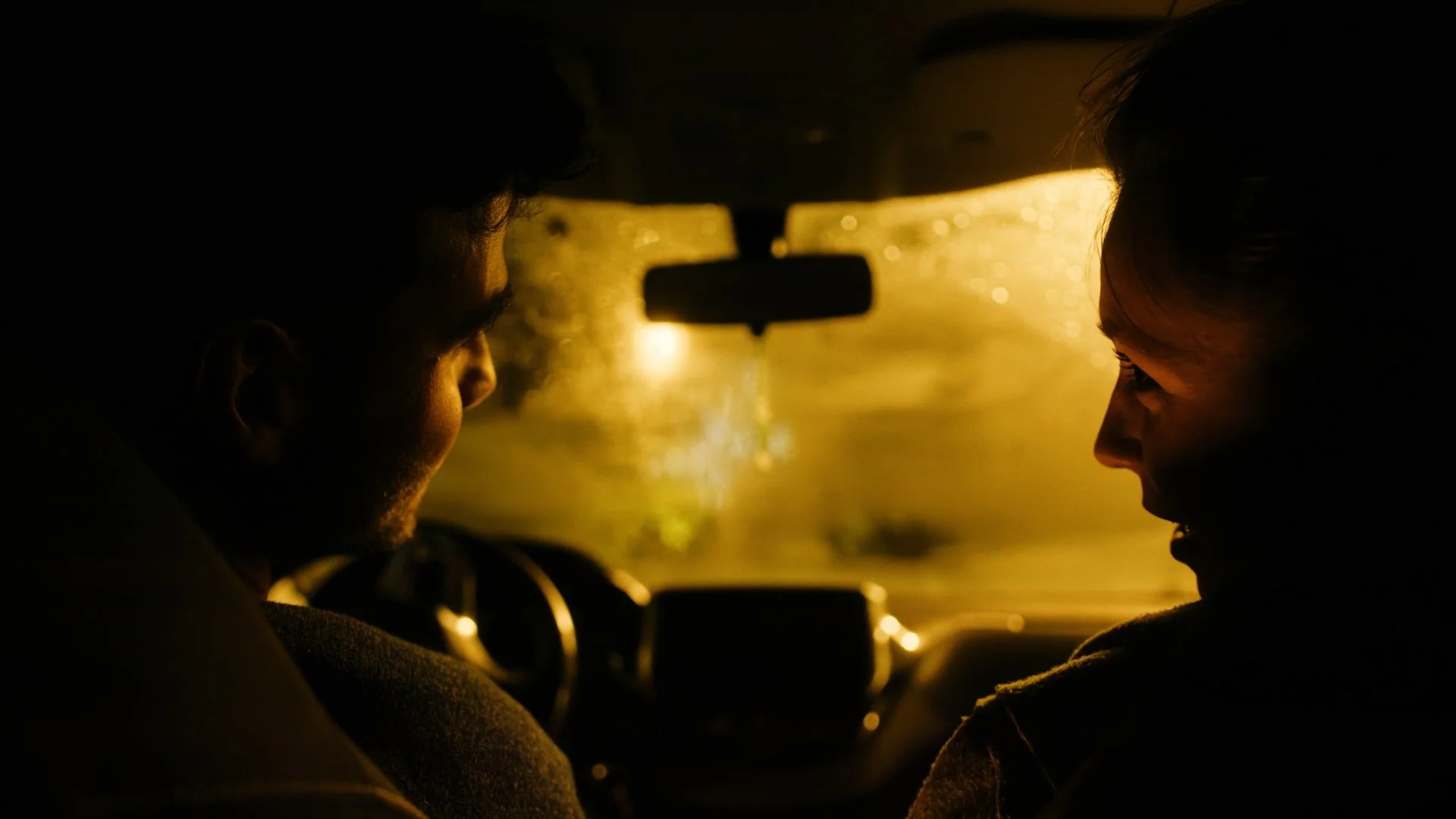 Two people facing each other inside a car at night, illuminated by yellow streetlights through a rain-streaked windshield