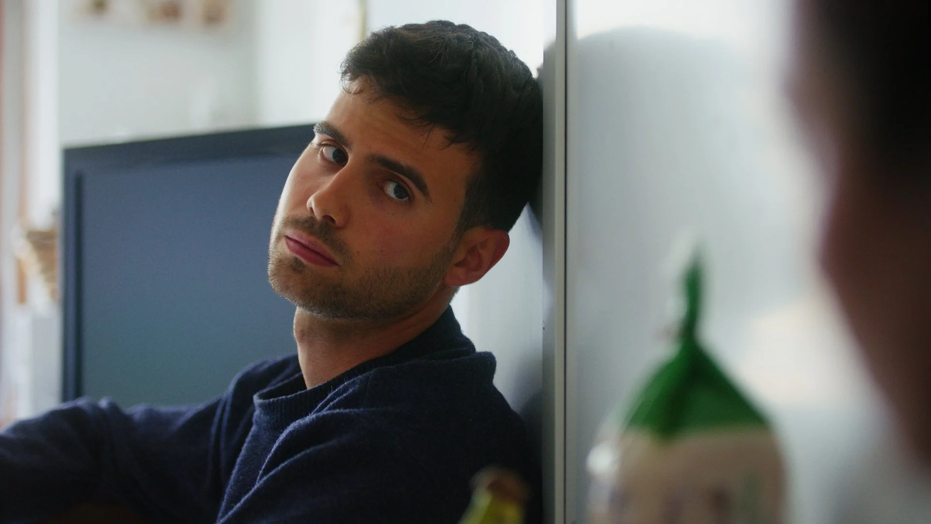 Man leaning against a wall indoors, looking towards the camera