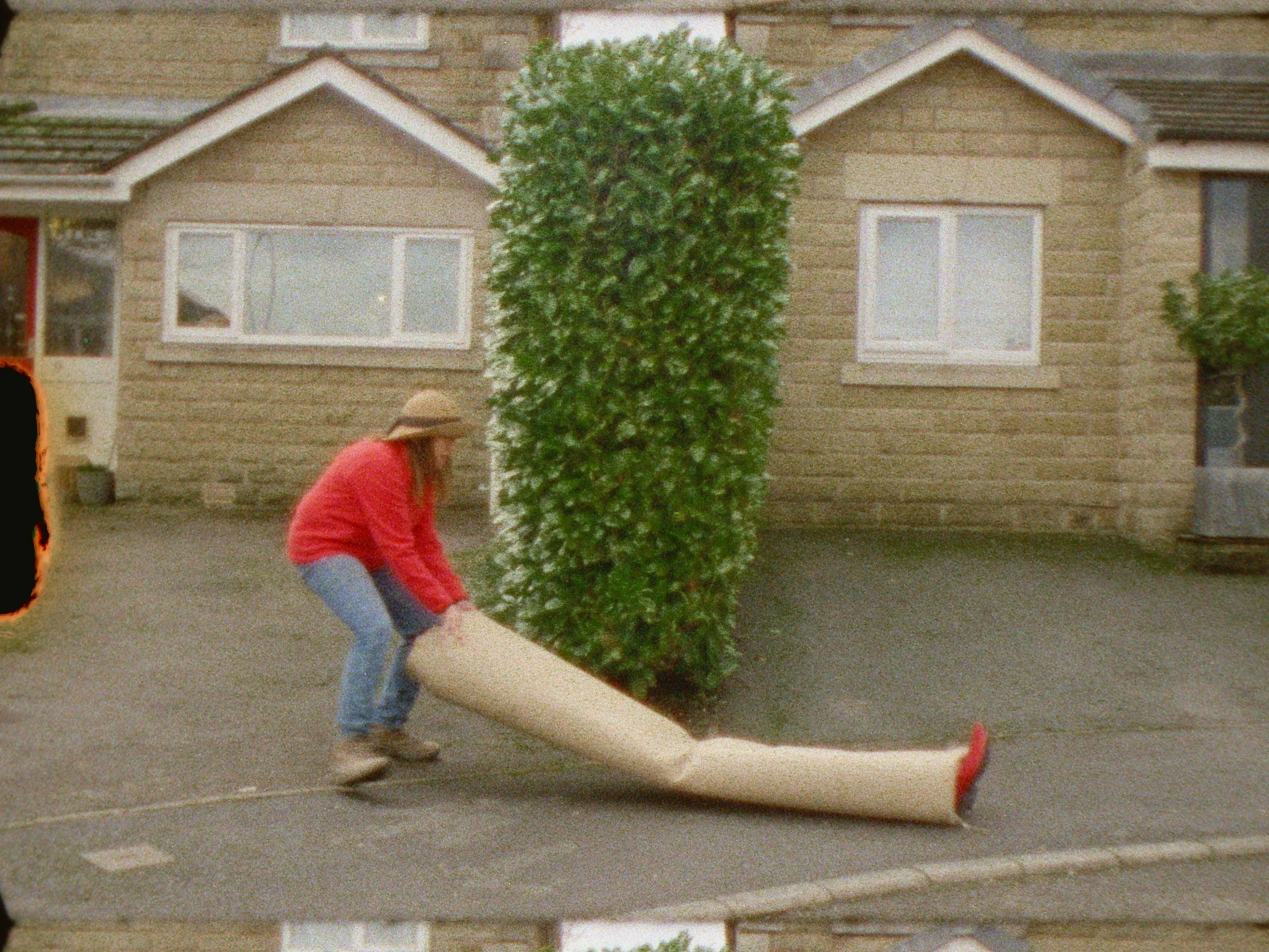 A person wearing a beige hat and red jacket is smoothing a large, rectangular piece of turf in a driveway in front of a brick house with white-trimmed windows