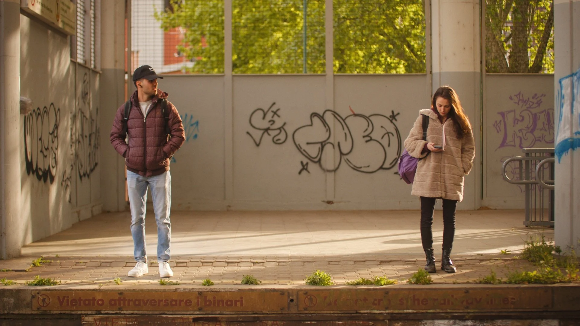 Man and woman standing outside at a train station, with graffiti and trees in the background