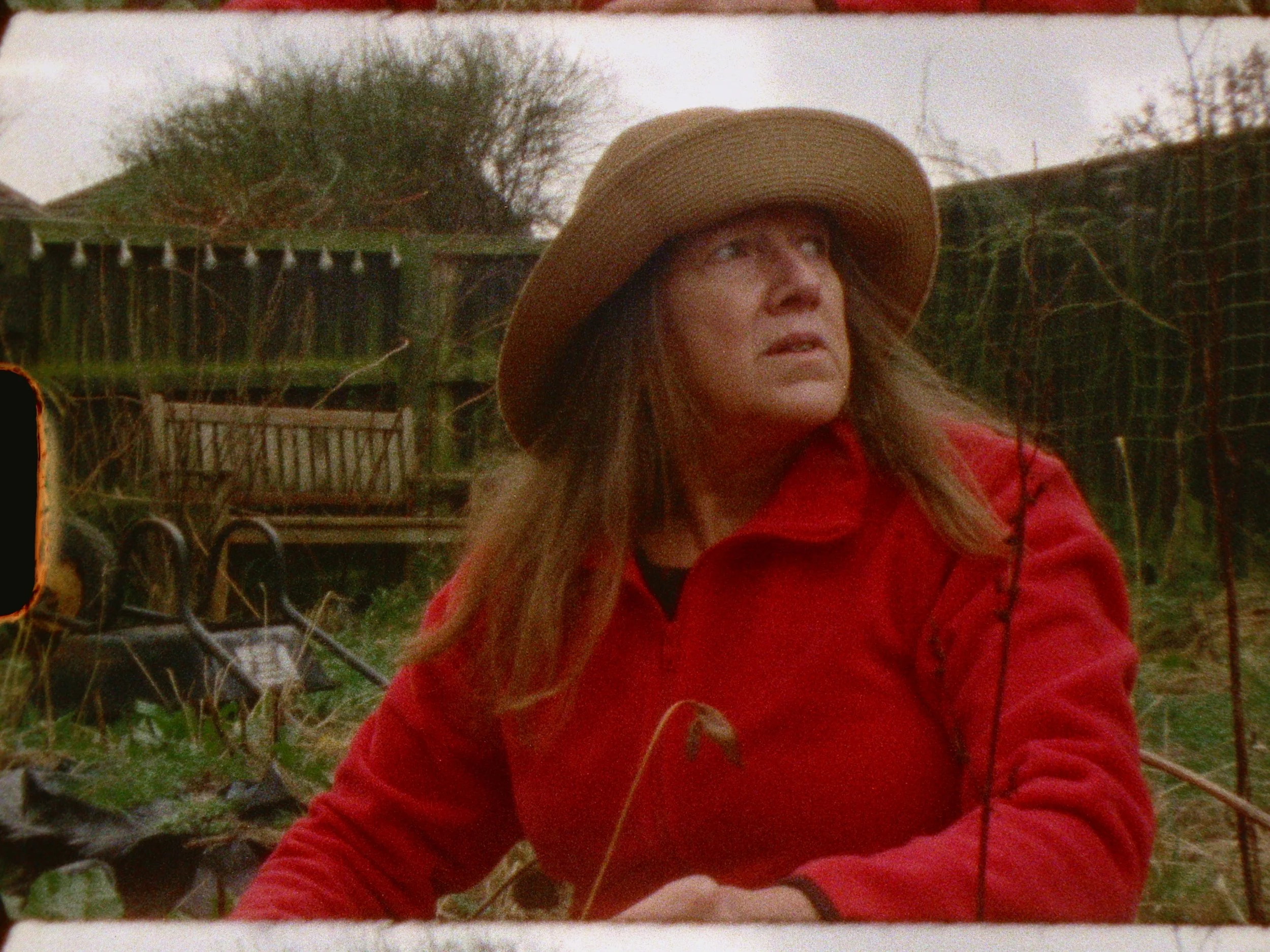A woman with long hair wearing a wide-brimmed hat and a red jacket, outdoors in a garden or yard with trees, a fence, and garden tools