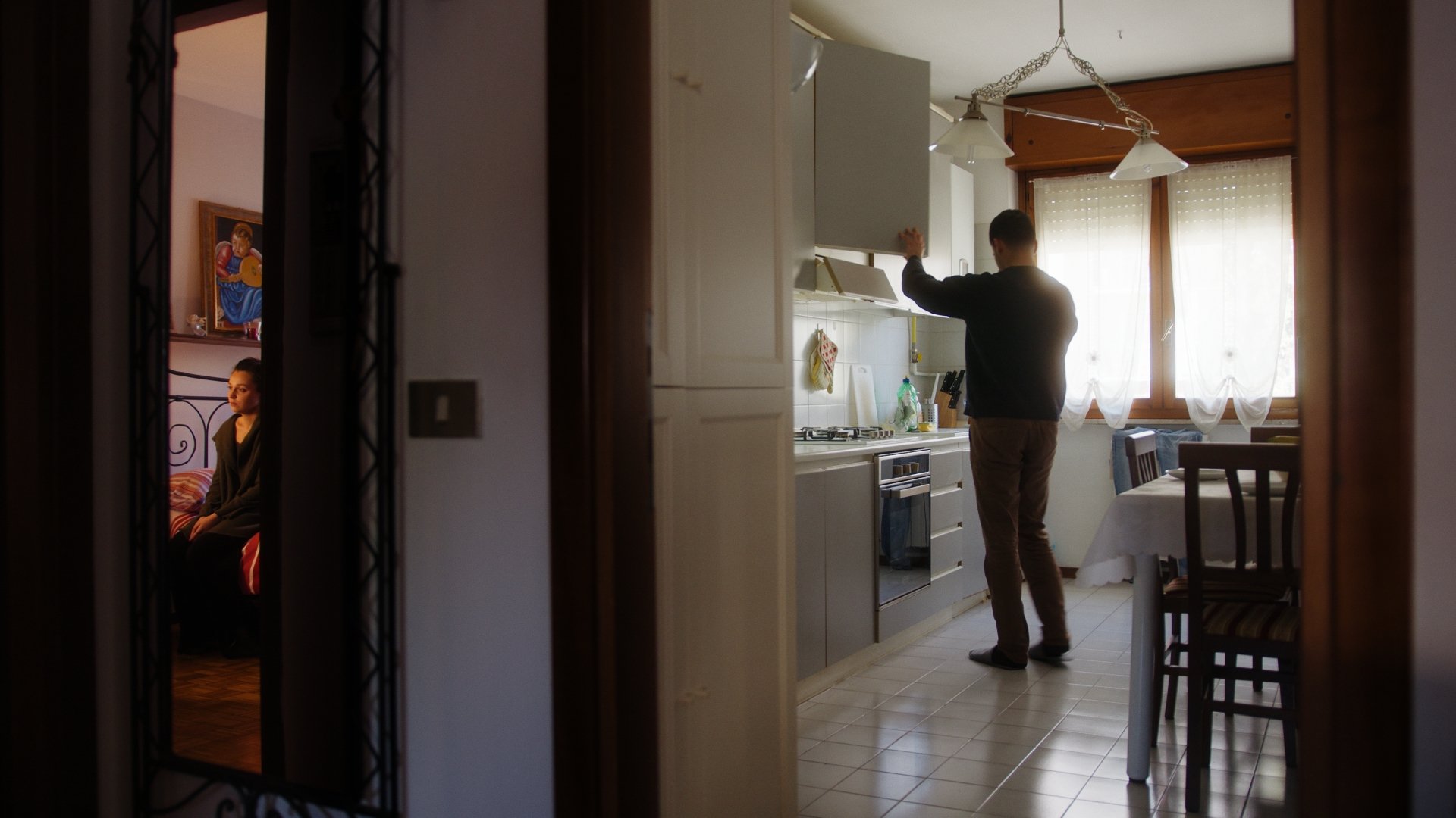 Person standing in a kitchen with sunlight coming through a window. A woman is sitting on a bed in a bedroom, seen through a doorway on the left side of the image