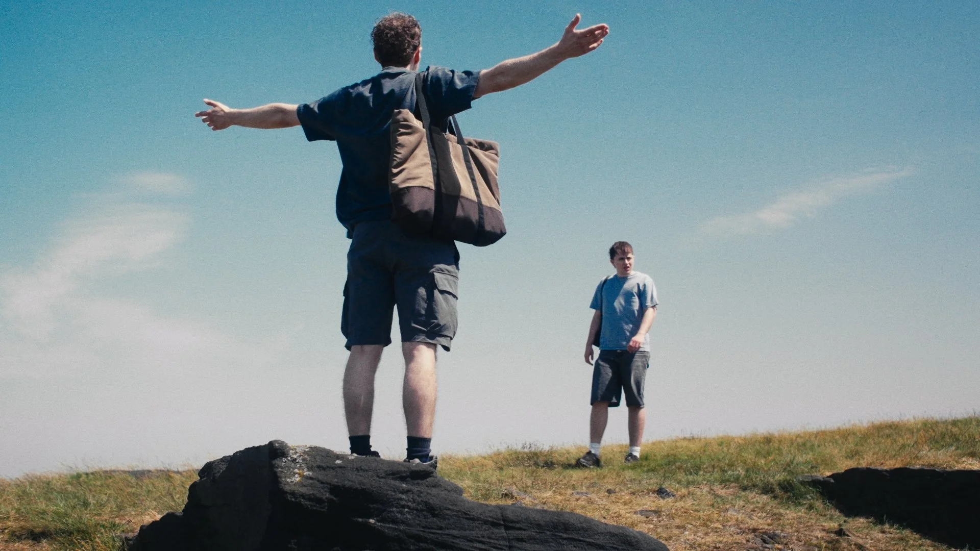Two young men standing outdoors on grassy terrain, one with arms outstretched and the other looking on