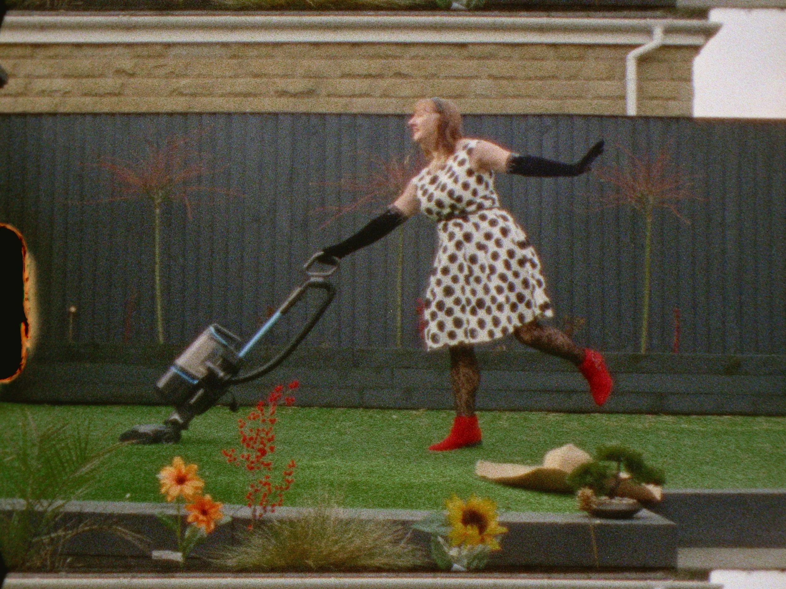 A woman dressed in a polka dot dress and red shoes, wearing black gloves, is holding a vacuum cleaner, vacuuming the grass