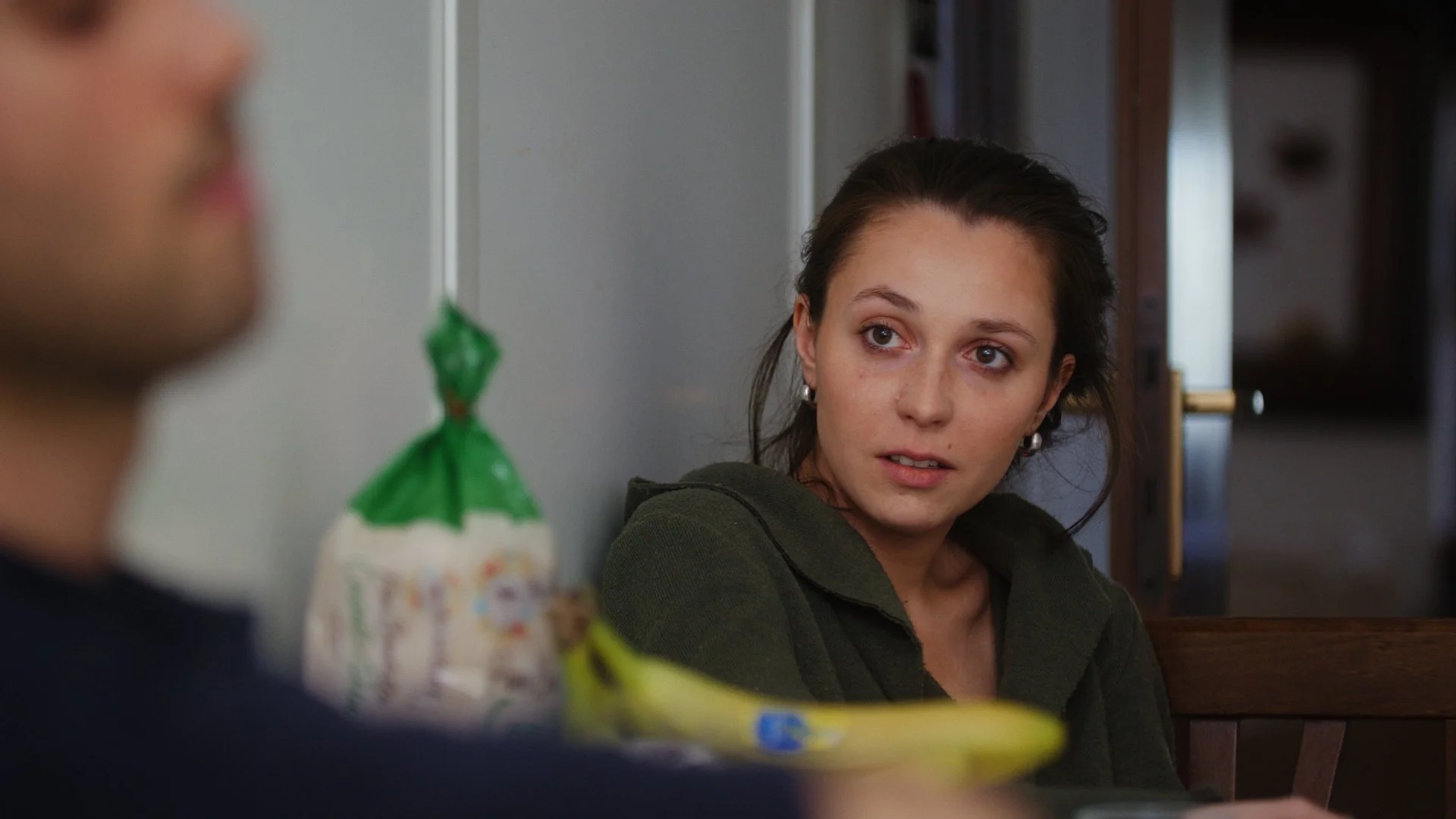 Woman in a green jacket sitting at a table, looking at a person out of focus in the foreground