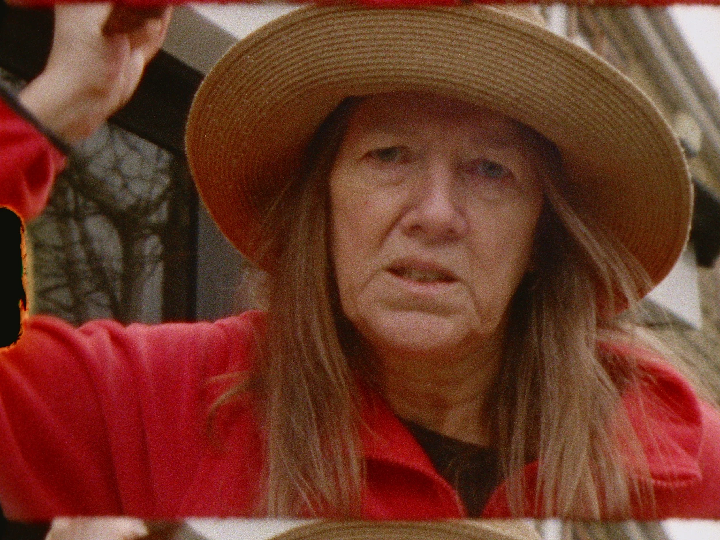 Close-up of a woman wearing a large straw hat and a red jacket,, stabbing the camera