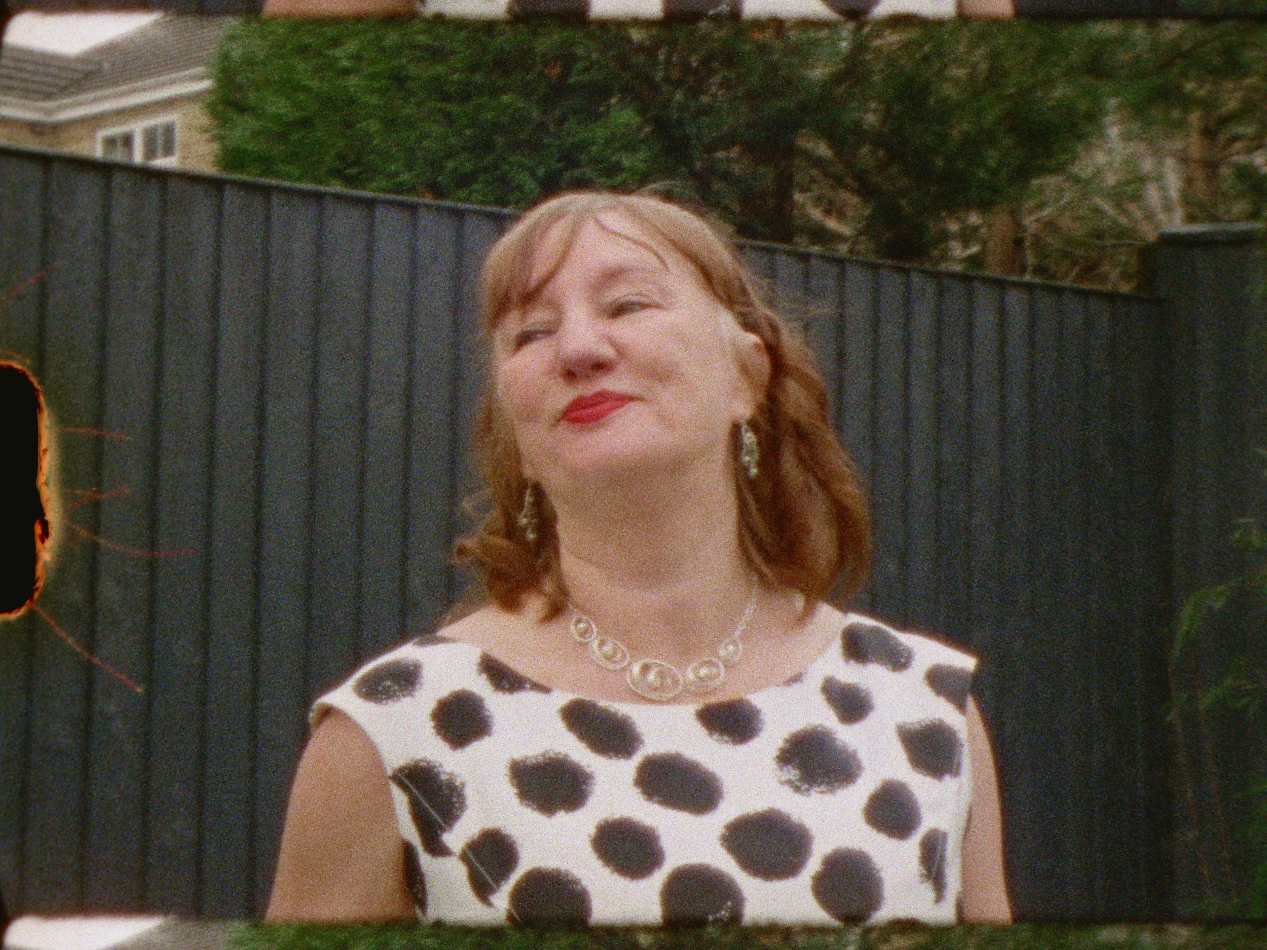 A woman with red hair and red lipstick smiling outdoors, wearing a white dress with black polka dots, a pearl necklace, and earrings