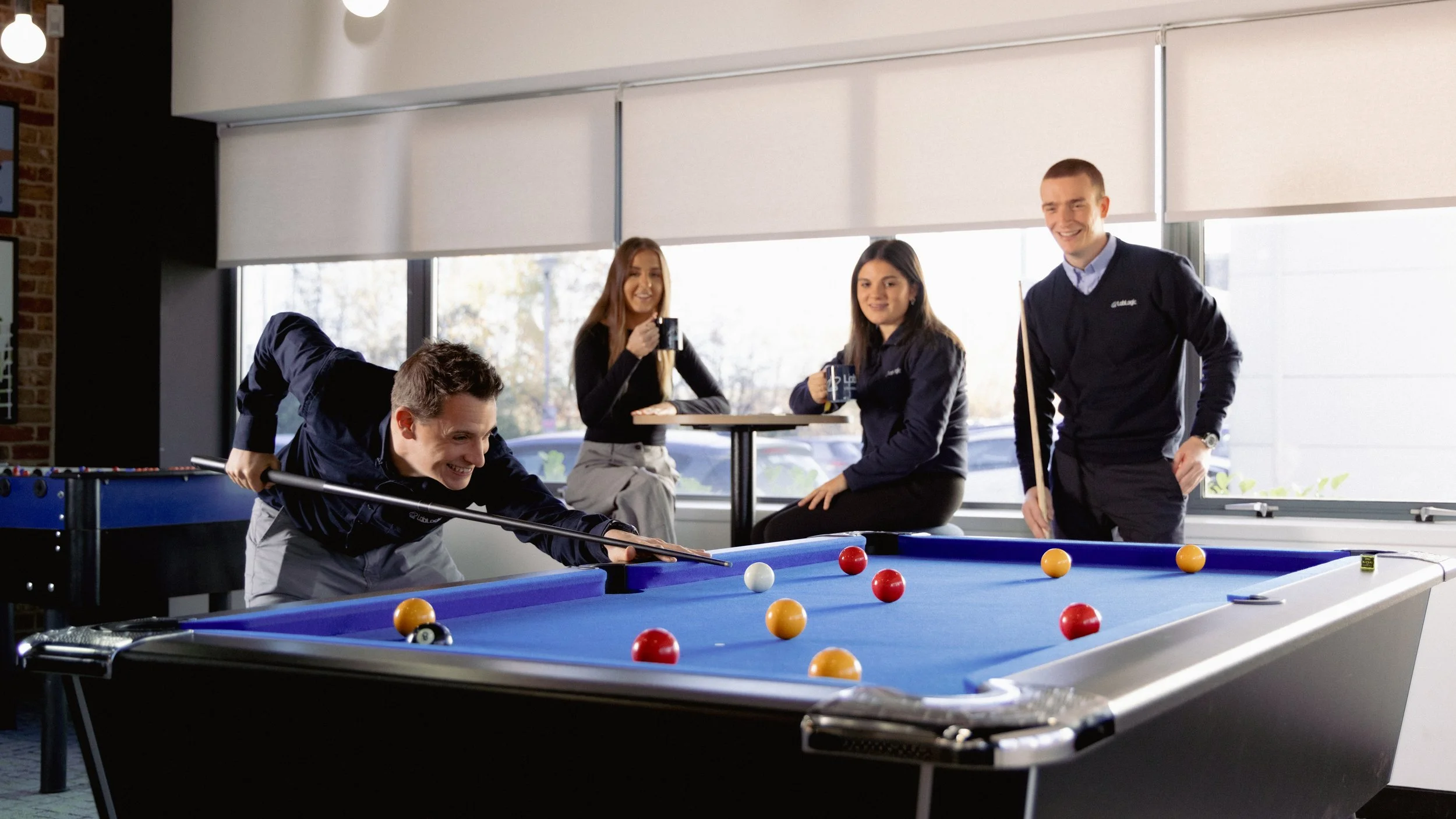 Four young adults in a room with large windows. One man is playing pool, leaning over the table with a cue stick. Three others, two women and one man, are sitting and standing with drinks, smiling and watching him.