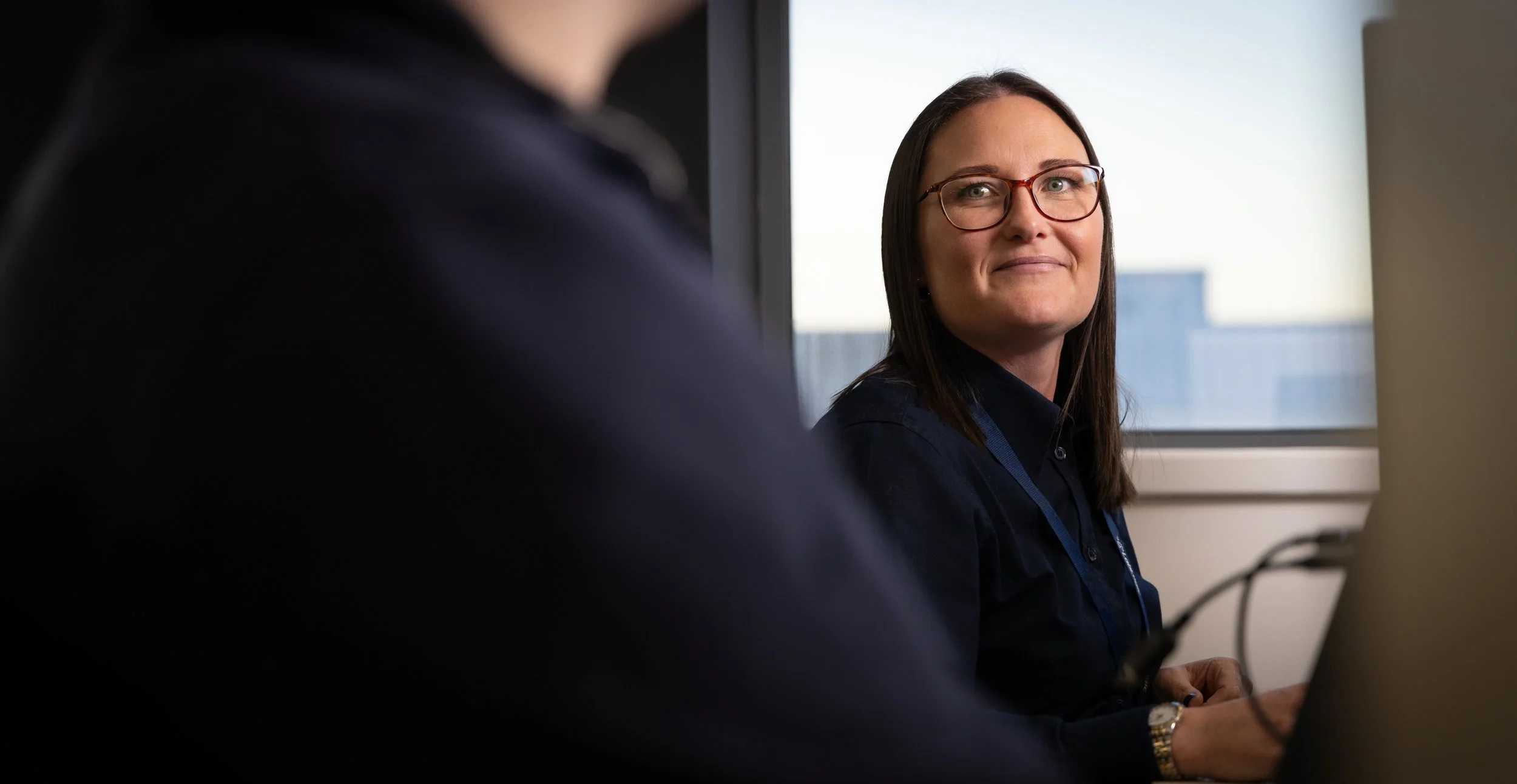 A woman with long brown hair and glasses sitting at a desk, looking at another person in front of her during a meeting in an office setting.