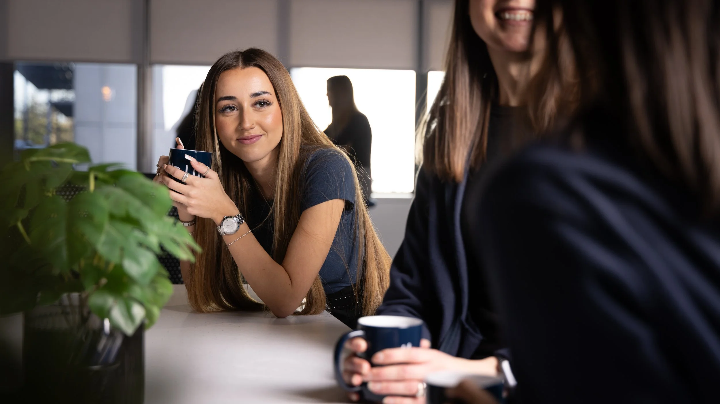A young woman with long brown hair sitting at a table, holding a coffee mug, smiling and looking at a person wearing a dark blazer, who is partially visible in the foreground. The background shows a modern office setting with large windows and other silhouettes of people.