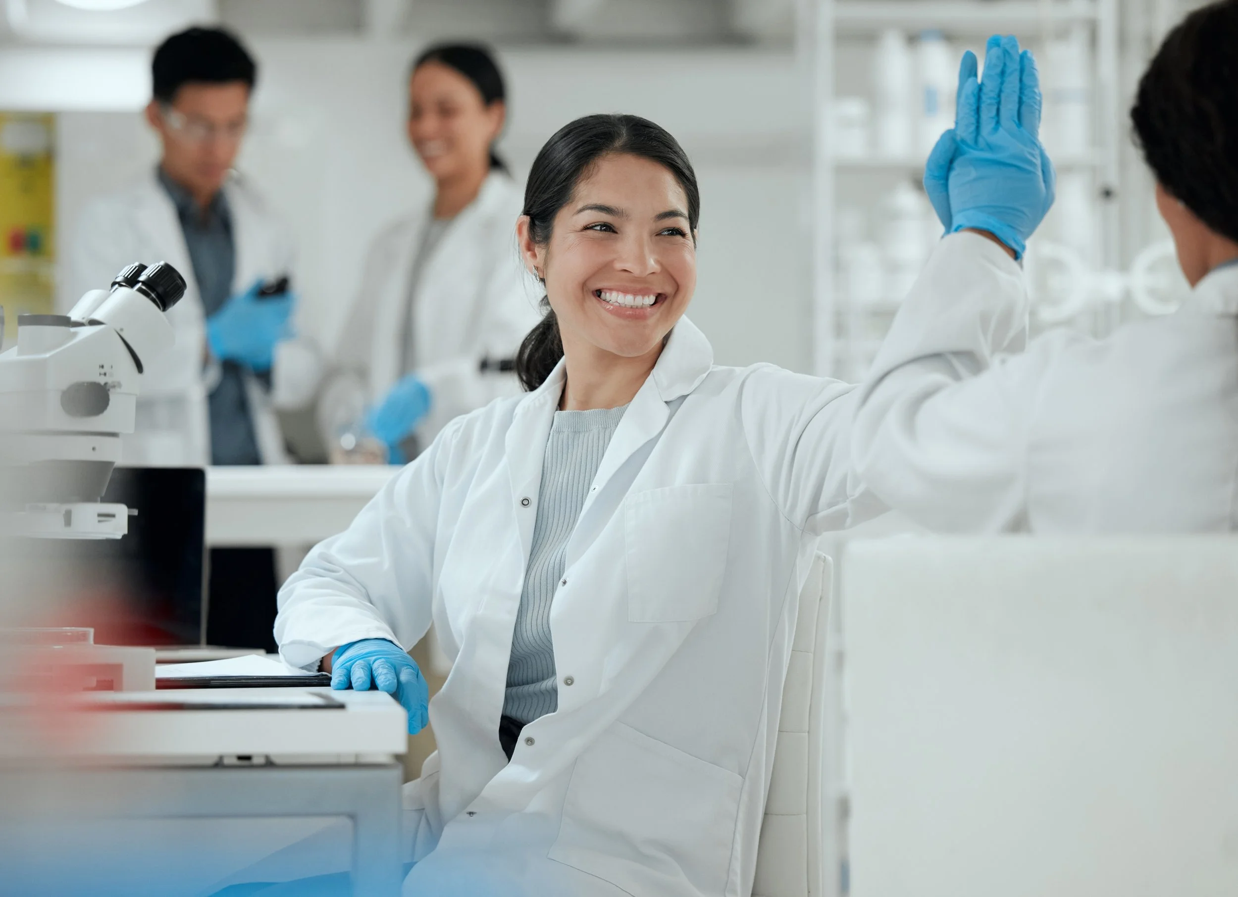 Scientists in a laboratory, with one woman smiling and giving a high five to a male colleague, both wearing white lab coats and blue gloves.