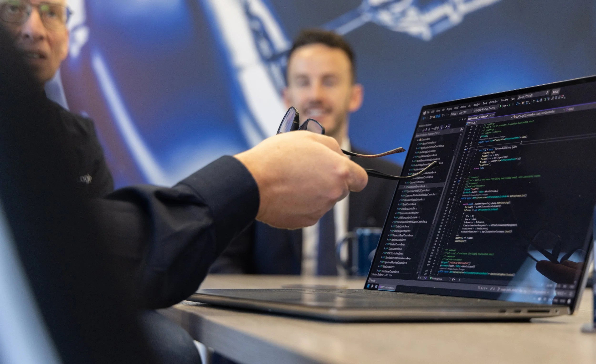 Close-up of a person holding eyeglasses in front of a computer monitor displaying programming code, with two individuals in a blurred background at a tech conference or office.