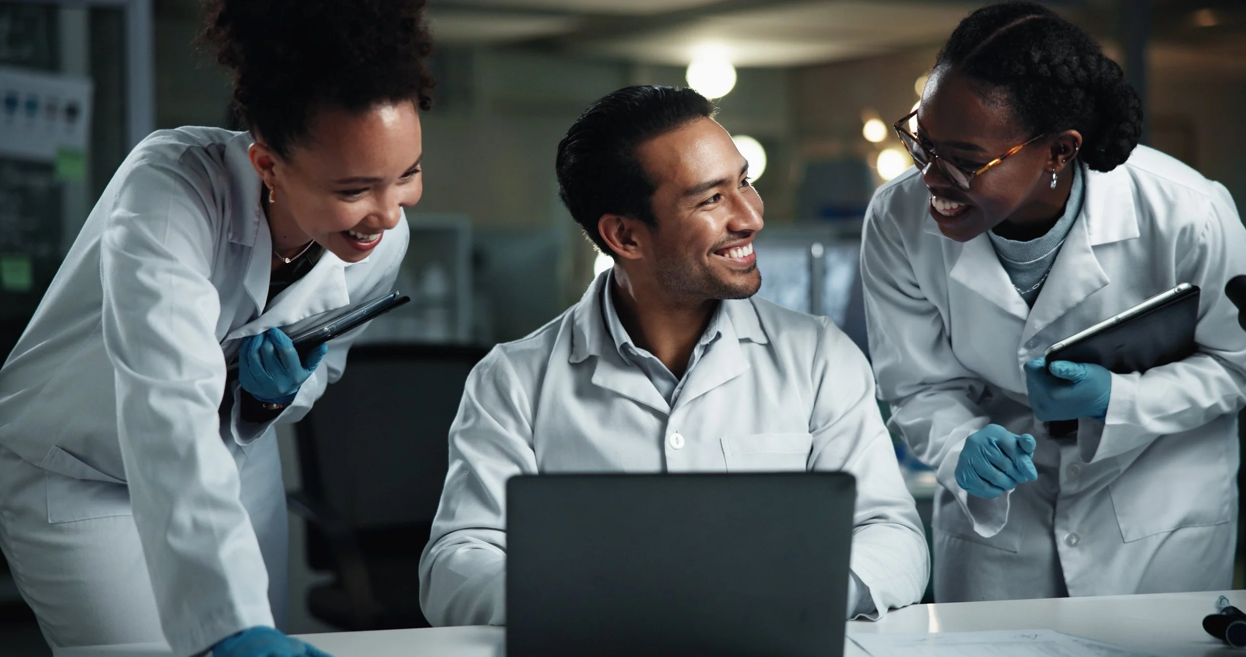 Three scientists in white lab coats are smiling and sharing a moment of joy while working together in a laboratory. One woman on the left is holding a tablet, another woman on the right is holding a tablet and wearing glasses, and a man in the center is sitting at a desk working on a laptop. All are wearing blue gloves.