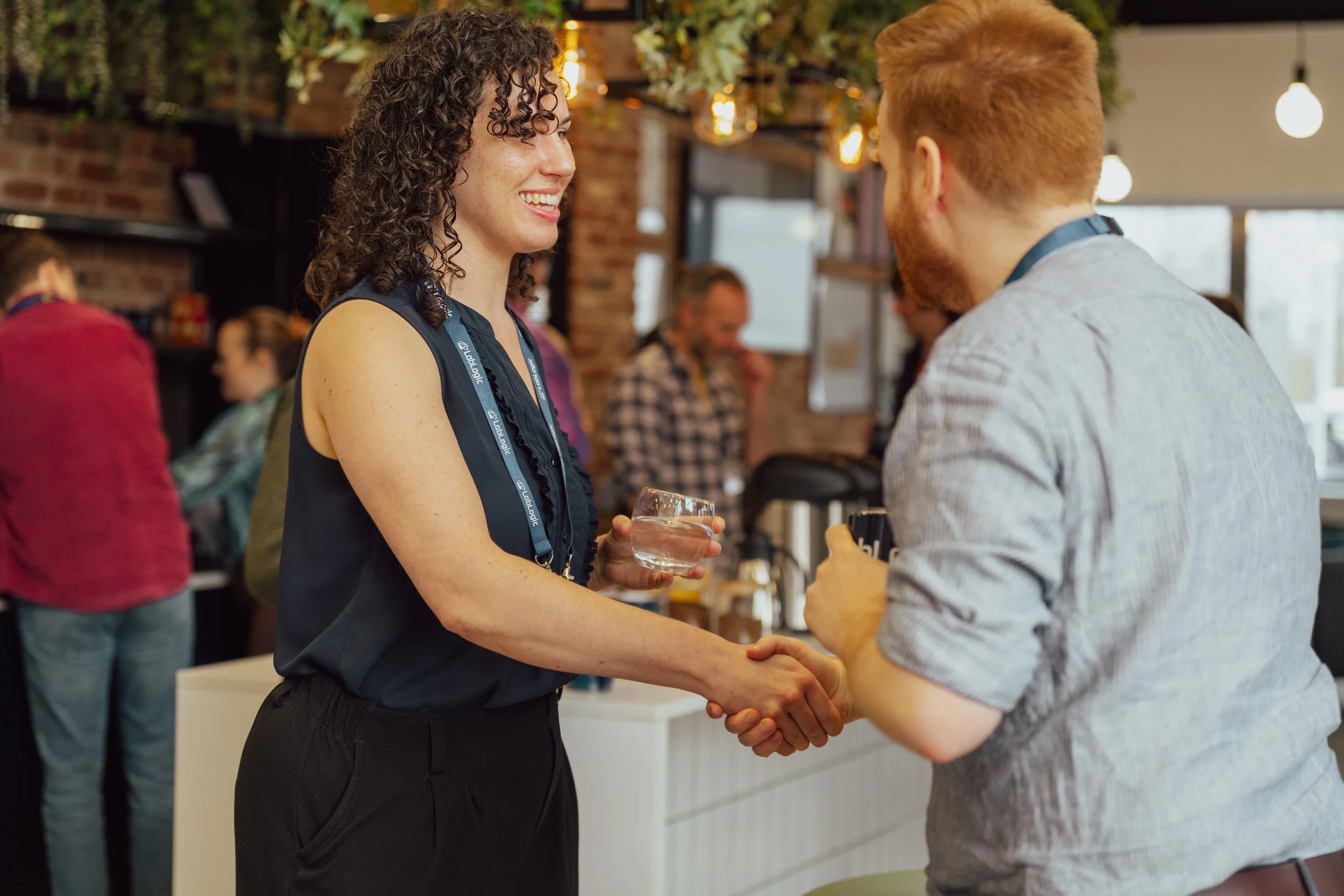 A woman with curly hair and a man with red hair shaking hands at a social gathering in a restaurant or café, with people in the background.