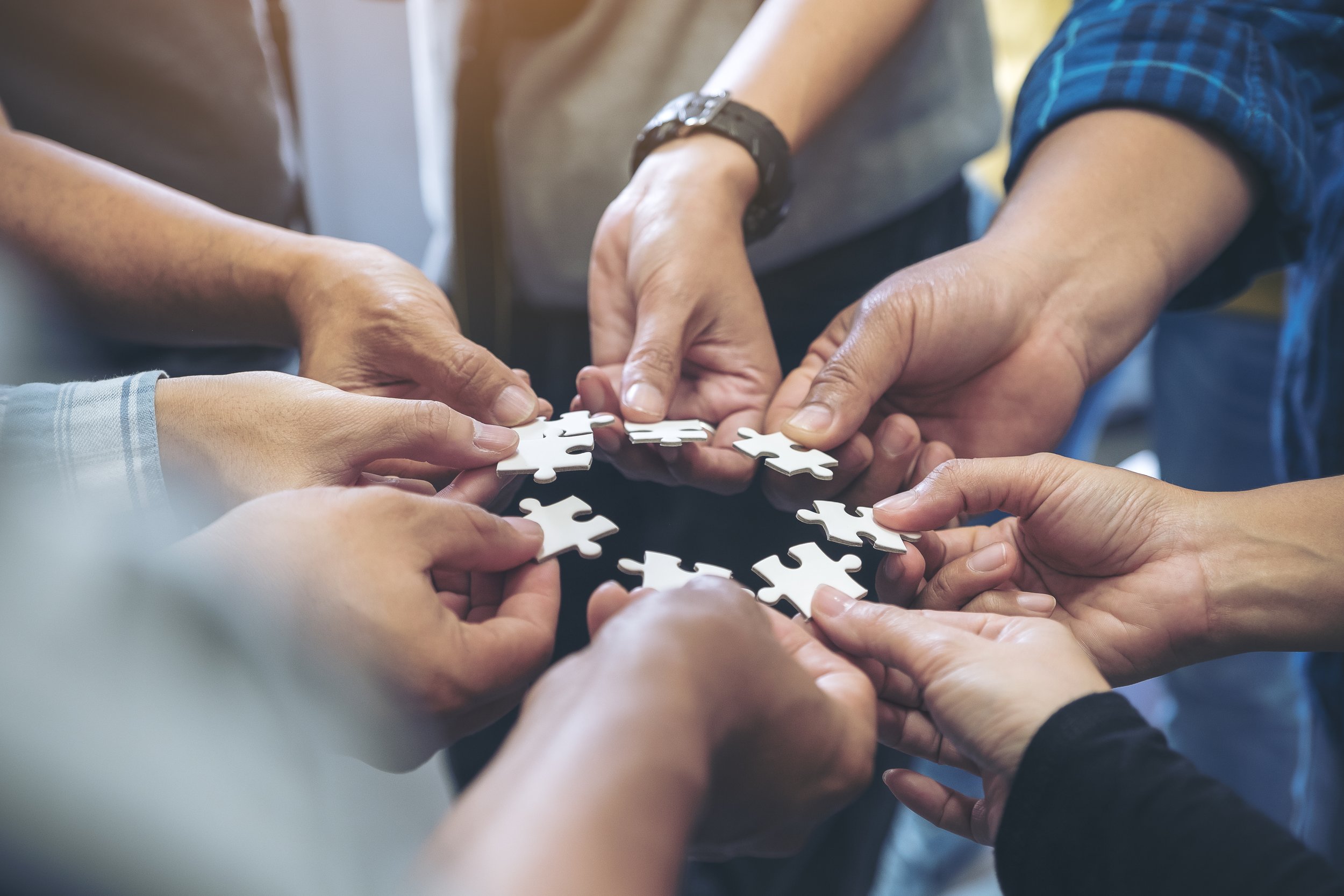 Multiple hands working together to assemble white jigsaw puzzle pieces.