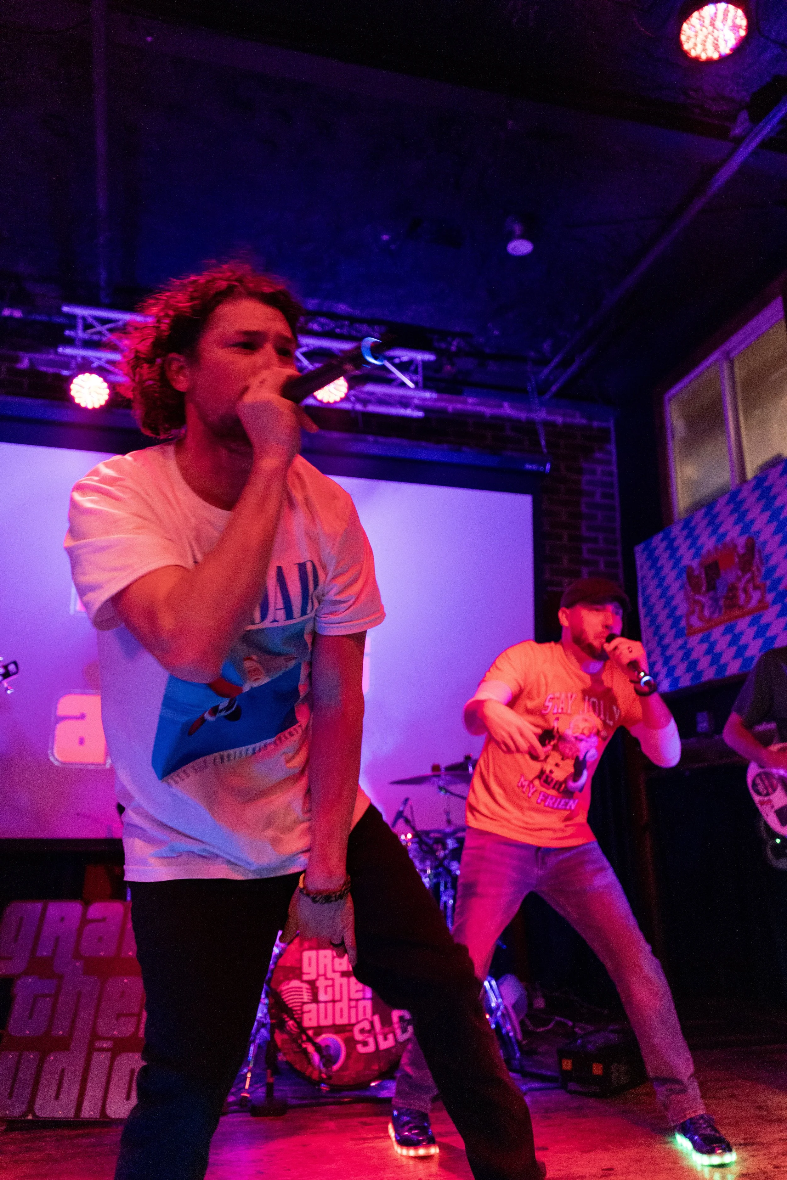 Two men performing on stage in a nightclub or concert setting with colorful purple and blue lighting. One man is singing into a microphone while the other is dancing, both wearing casual T-shirts. The background has music equipment and a screen.