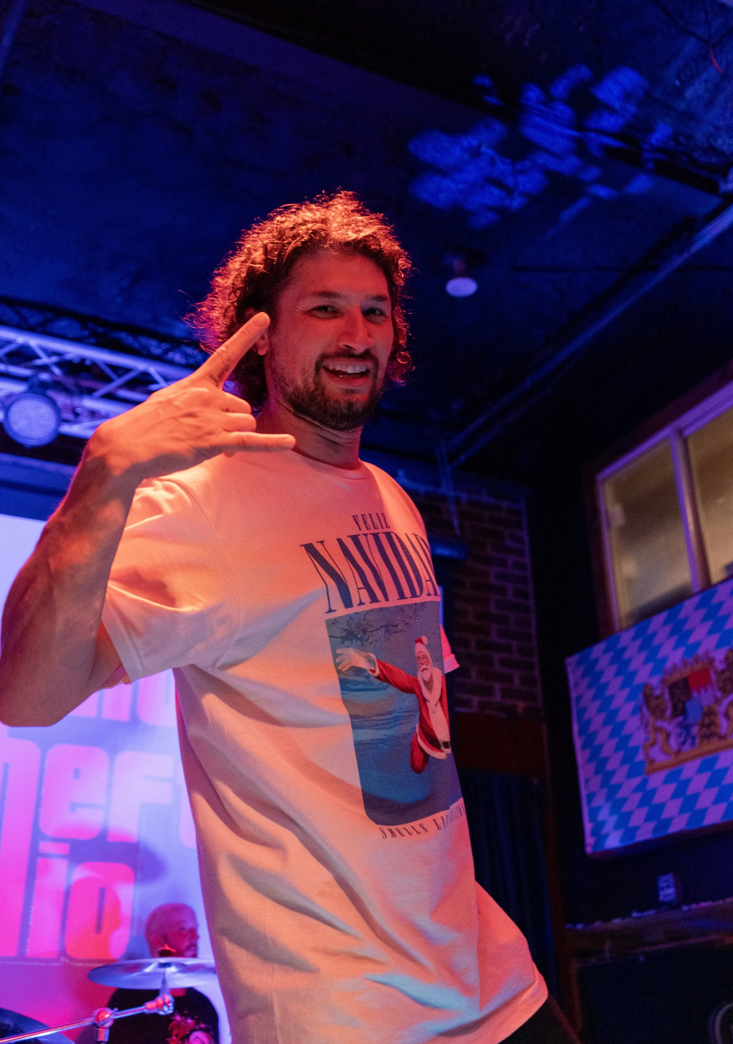 A man with curly hair and facial hair making a rock hand gesture while smiling, wearing a graphic T-shirt in a dimly lit room with purple and blue lighting, and a musician playing drums in the background.