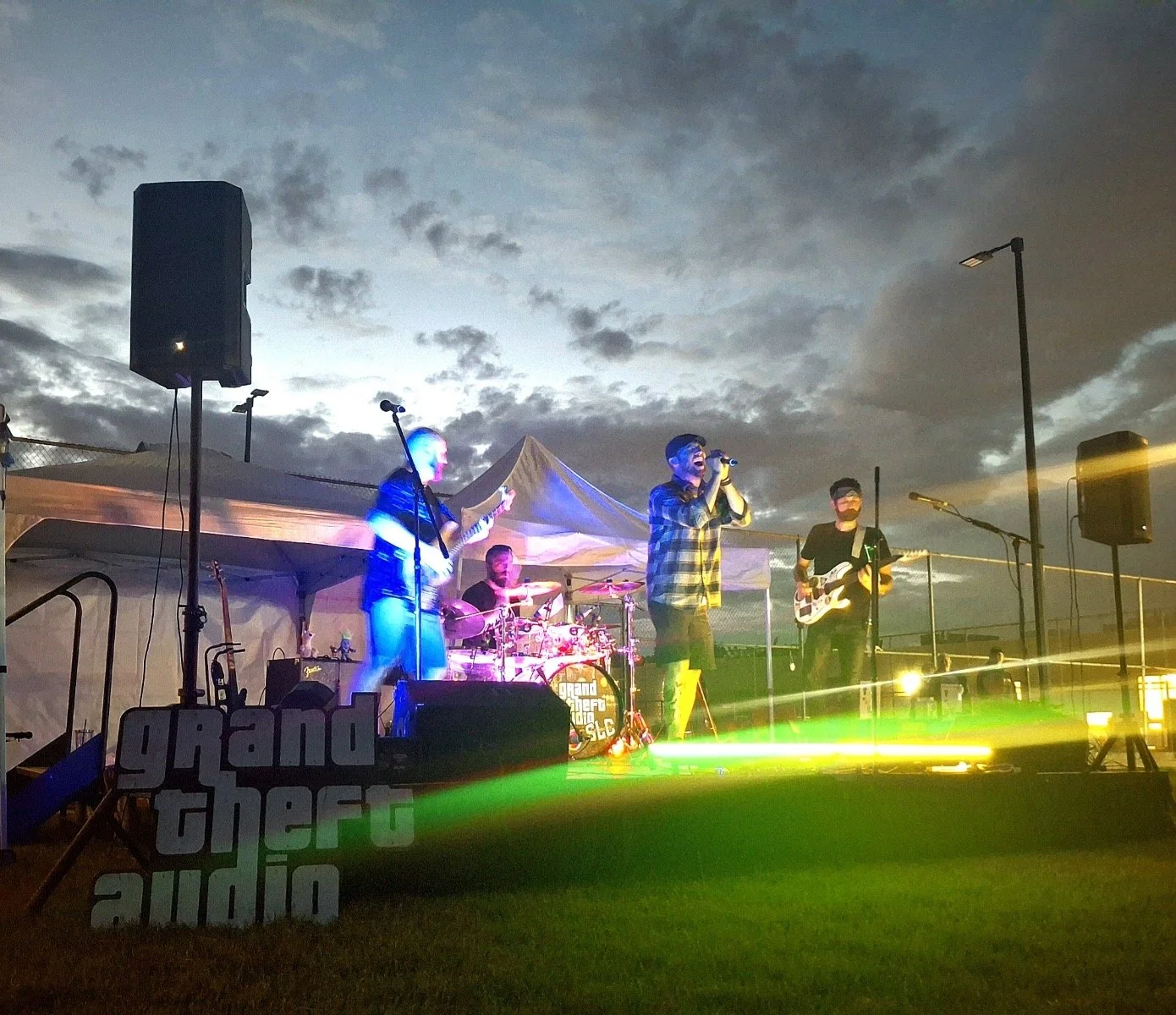 A band performing live on an outdoor stage at dusk, with four members: one singing into a microphone, two playing guitars, and one on drums. The stage is set up with a tent, large speakers, and the words 'grand the traudio' displayed on the front. The sky above is cloudy and the scene is illuminated with colorful lights.