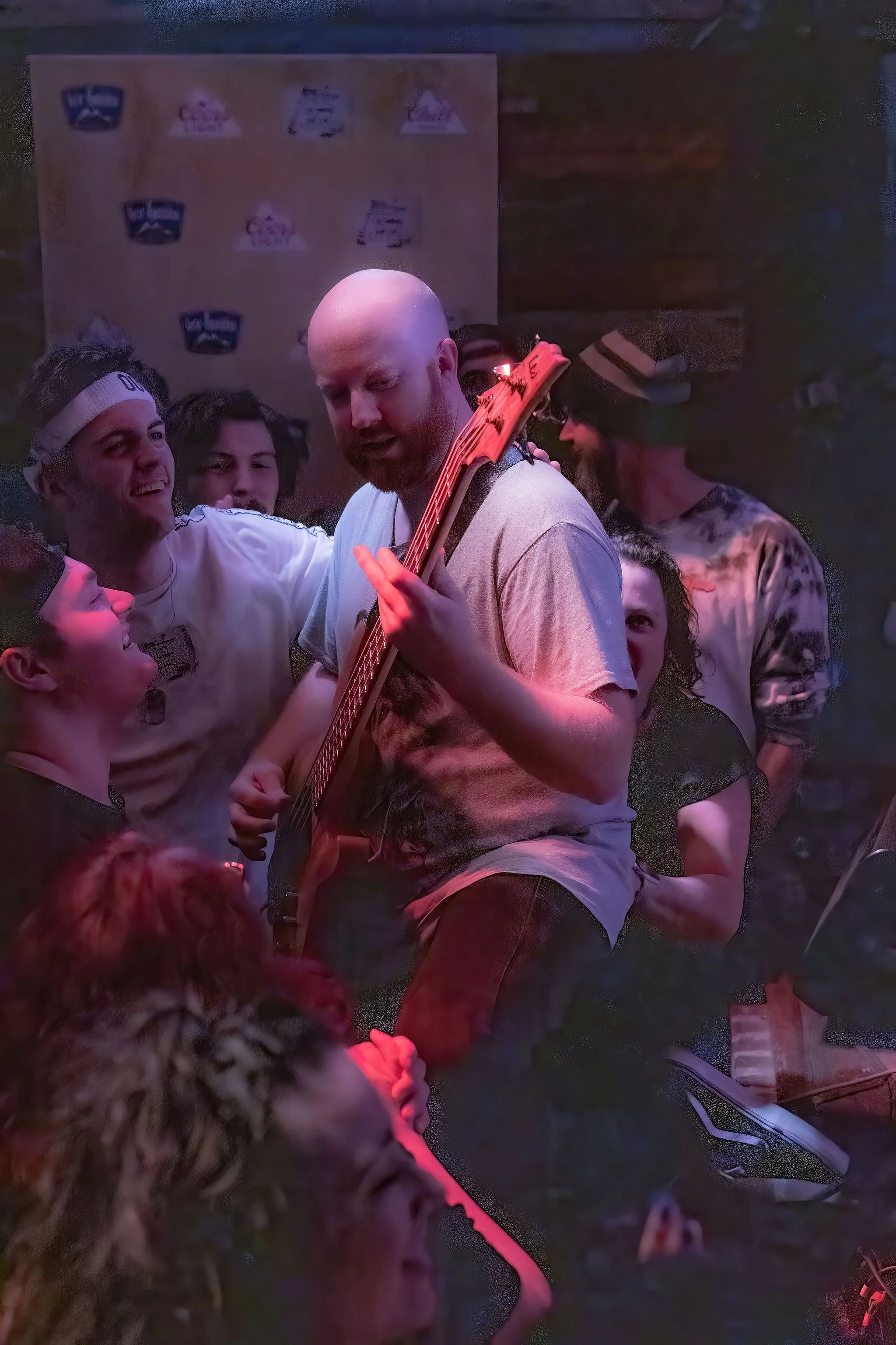 Group of young people, including a man playing electric guitar, gathered together in a dimly-lit room with posters on the wall, enjoying a social moment.