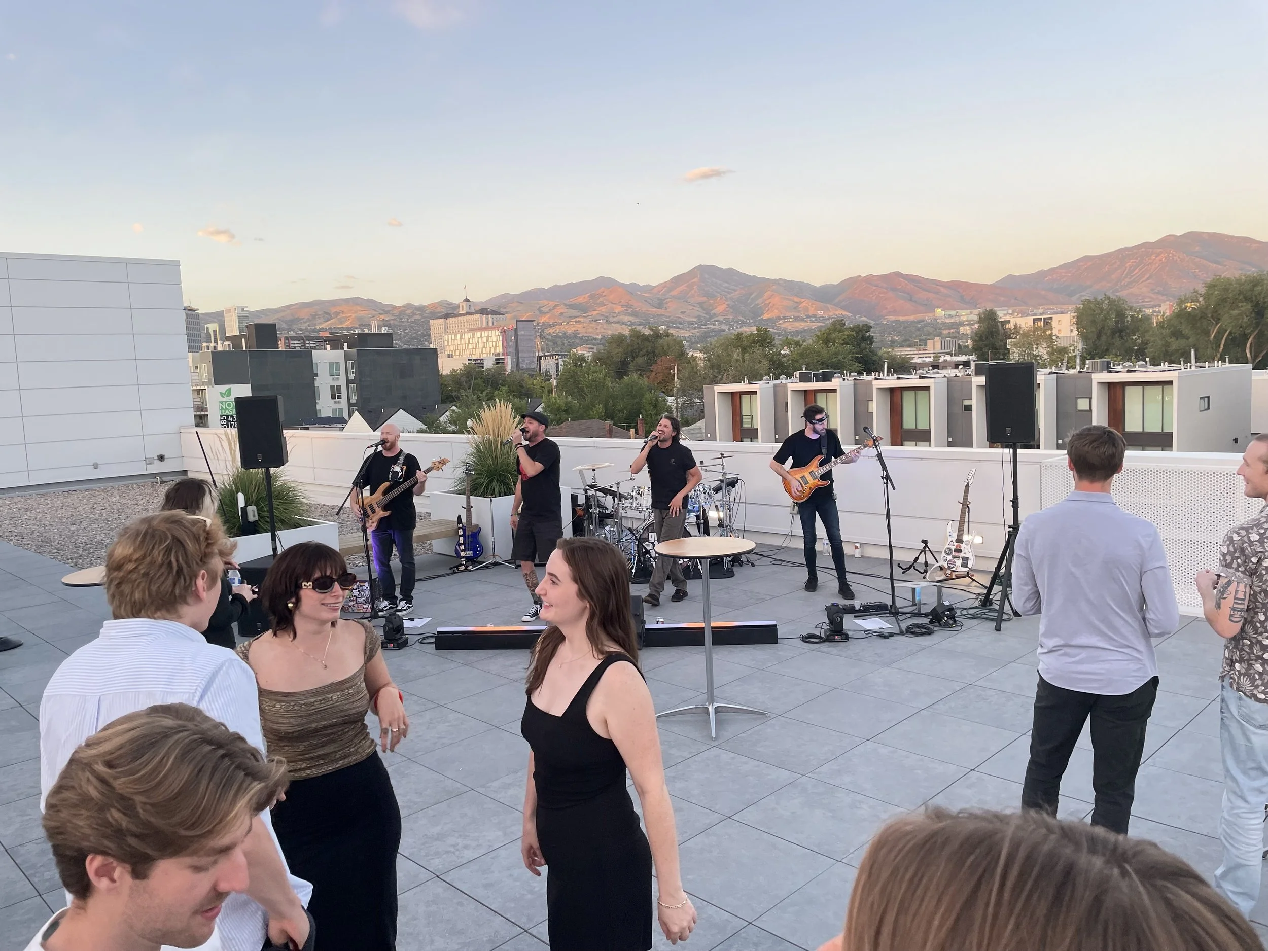 A rooftop party with a live band performing, guests dancing, and a scenic mountain view at sunset.